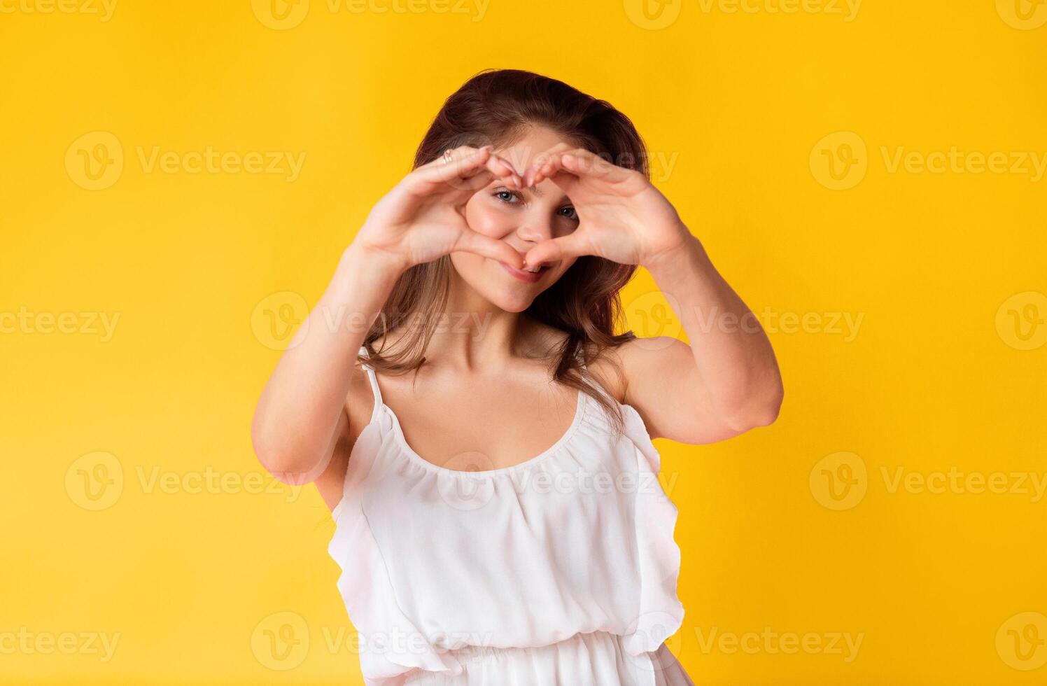 A young woman is playfully forming a heart shape with her hands. She stands confidently in front of a vibrant yellow backdrop, wearing a light-colored dress that adds to her cheerful demeanor. photo