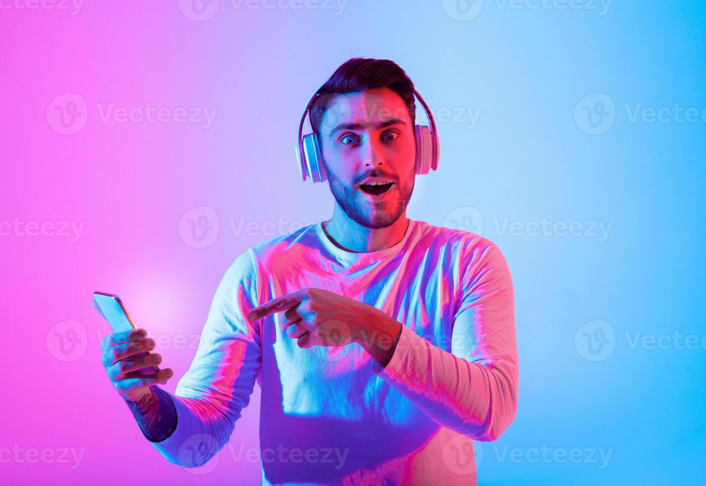 A man with headphones smiles while pointing at his smartphone illuminated by colorful lights. His expression reflects excitement and engagement with the music he is enjoying. photo