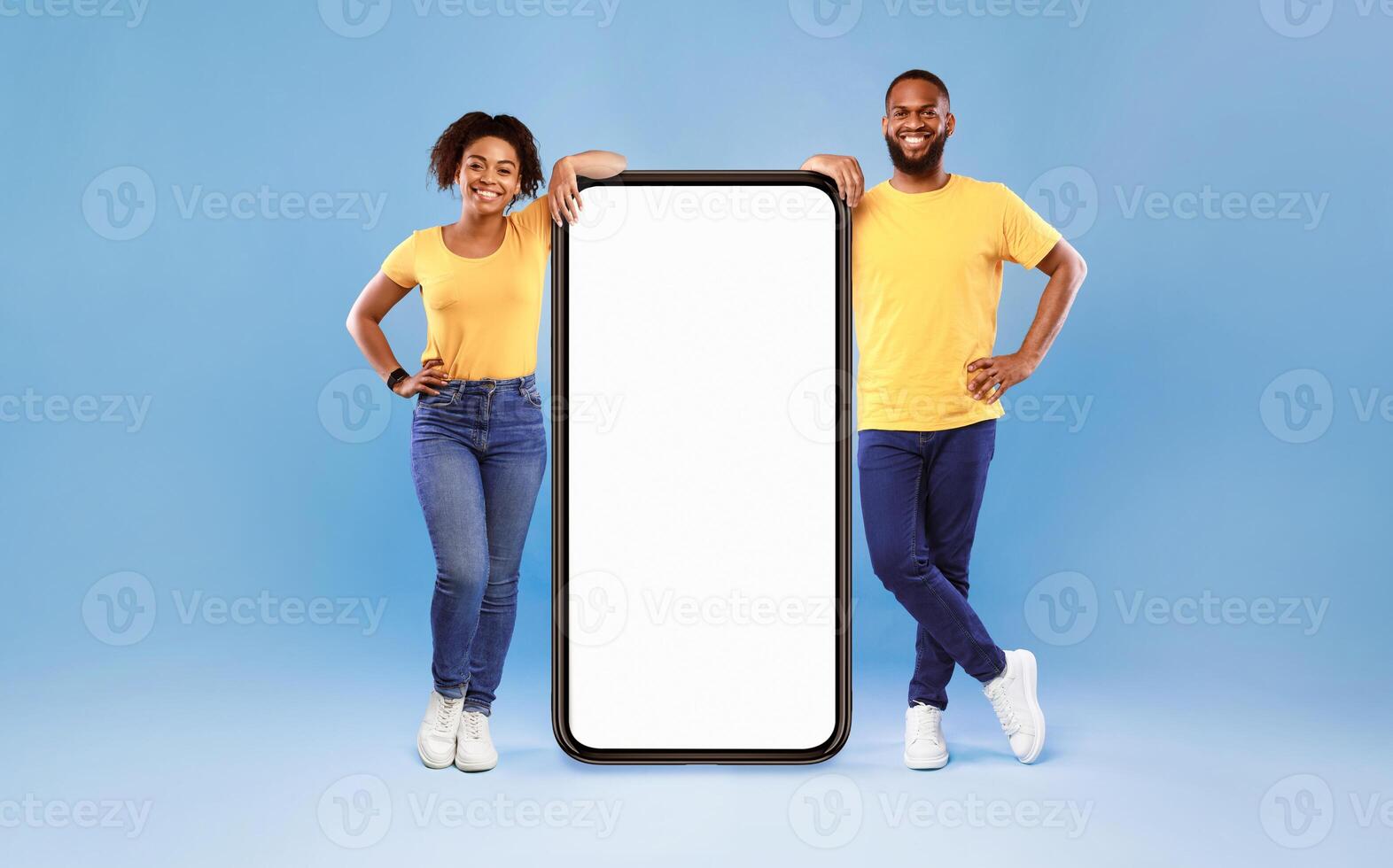 A joyful African American couple poses together, smiling while leaning on a large smartphone. They wear matching yellow shirts and casual outfits, highlighting a modern lifestyle. photo