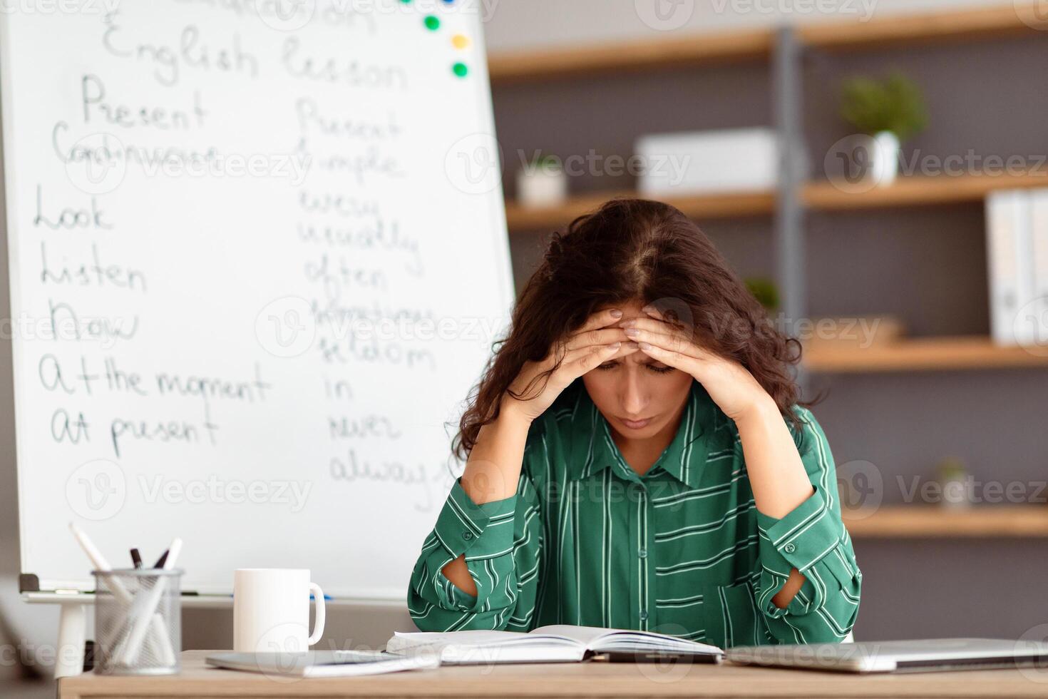 A young Arab woman sits at her desk, visibly stressed while studying lesson plans for remote teaching. She is surrounded by educational materials and a whiteboard filled with notes. photo