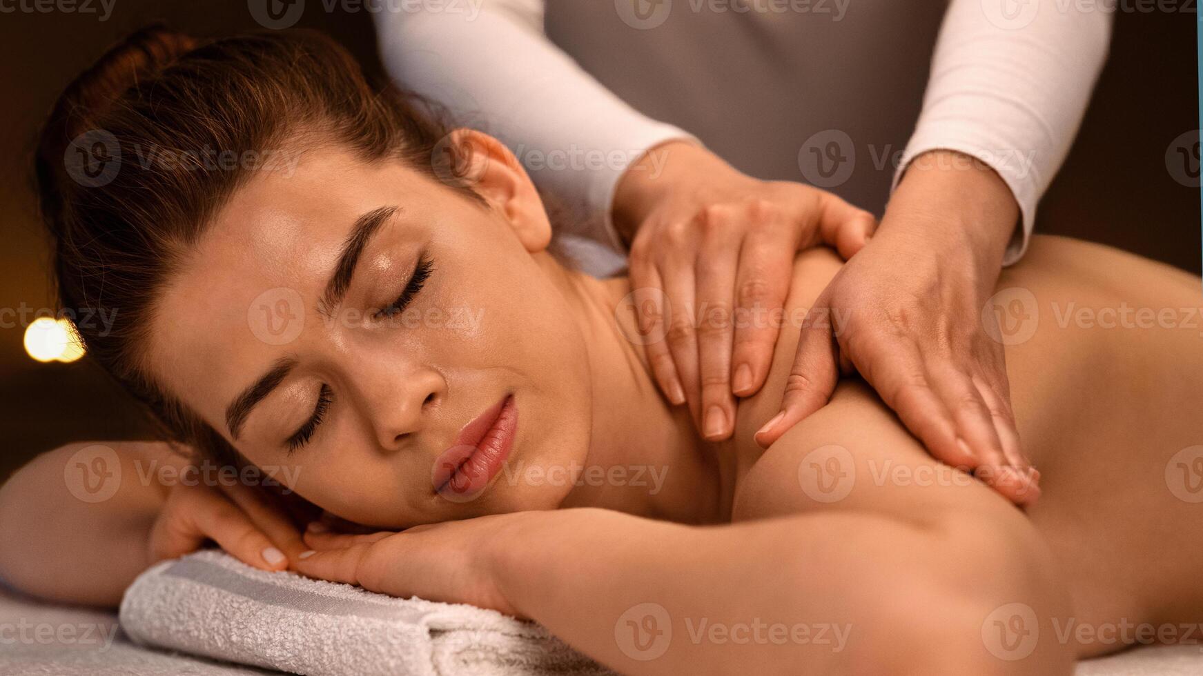 Relaxed young woman having healing body massage at newest luxury spa decorated with lightened candles. Female therapist rubbing sleeping lady back, giving her relaxing massage, side view, closeup photo
