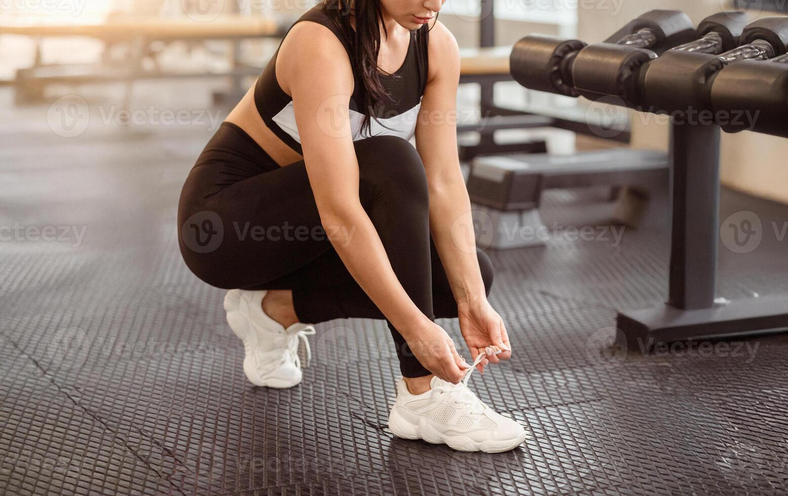 A woman in athletic clothing squats down to tie her shoelaces while preparing for a workout at a gym. The setting features dumbbells and exercise equipment in the background. photo