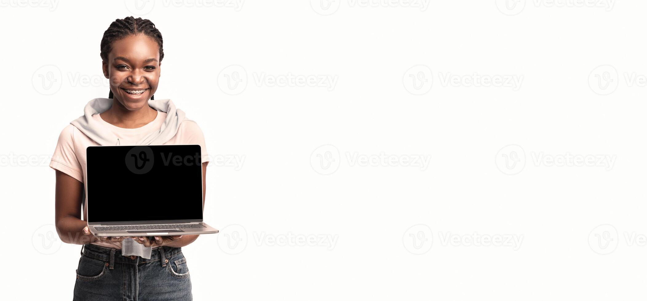 A cheerful young woman stands confidently, holding a laptop in her hands. She is wearing casual clothes and showcases a bright, welcoming smile against a neutral backdrop. photo