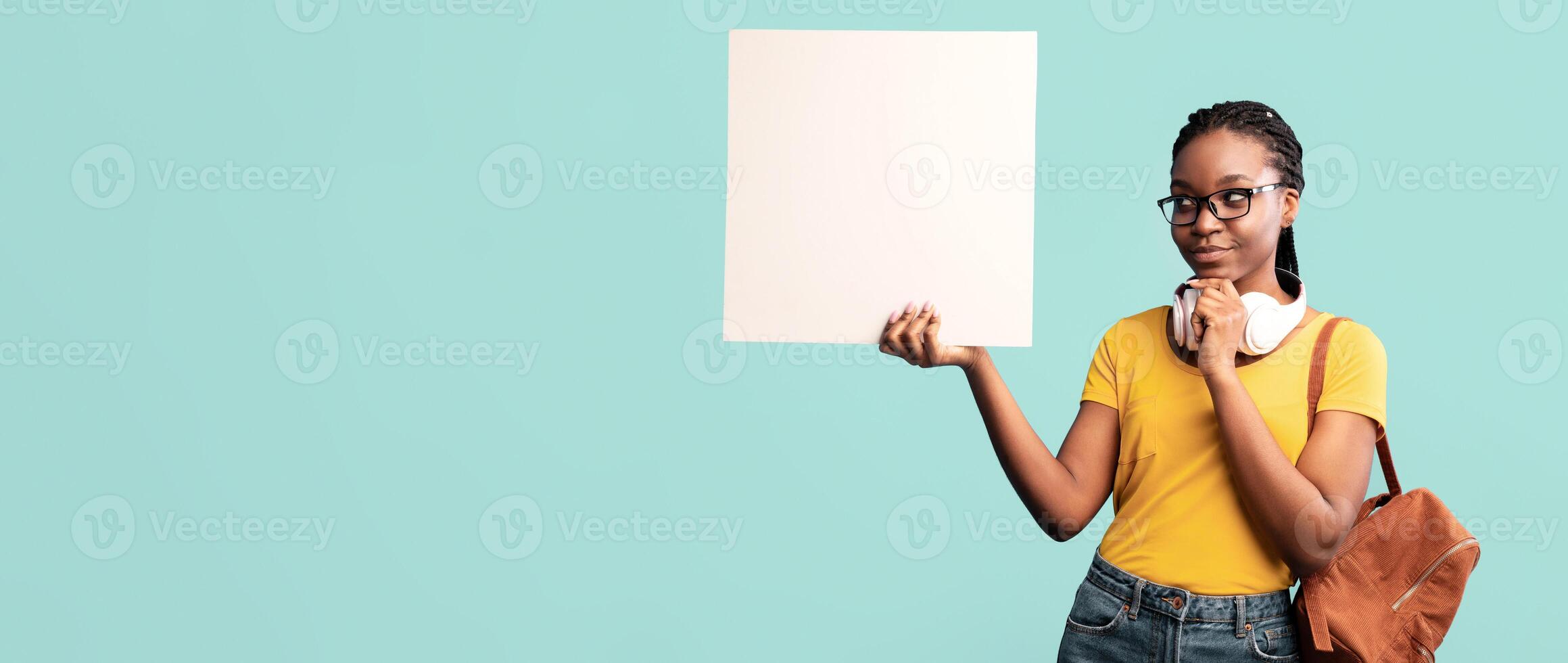 A young woman stands in front of a light blue wall, holding a blank canvas with one hand while thoughtfully pondering with a smile. She wears a yellow shirt and denim shorts. photo
