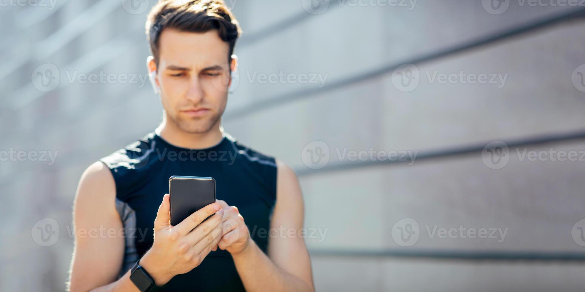 Read sports blog or choose a song. Serious guy in sportswear with wireless headphones typing at smartphone on gray wall background, free space, sun flare photo