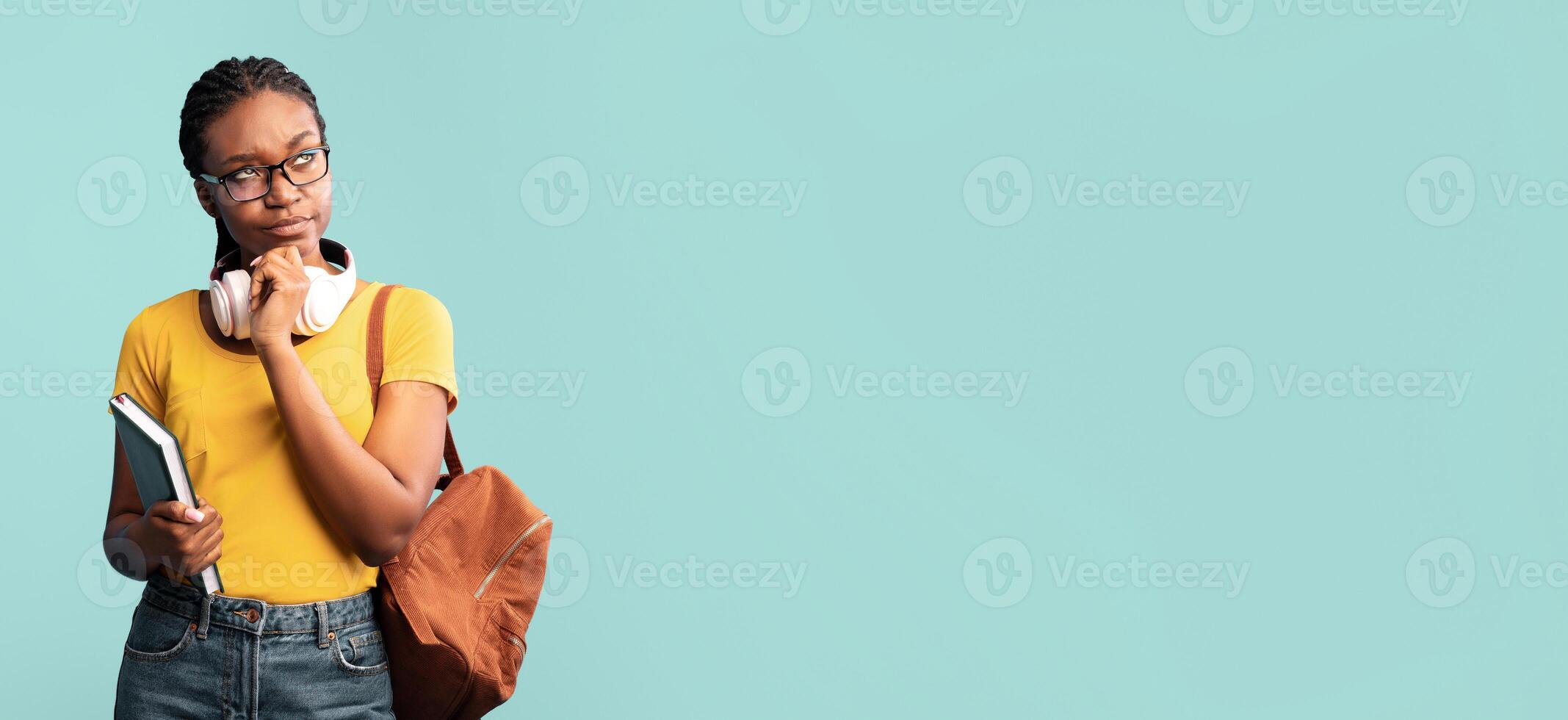 A thoughtful young student stands against a solid teal backdrop while holding a notebook. Wearing headphones around her neck, she appears deep in contemplation, perhaps preparing for studies. photo