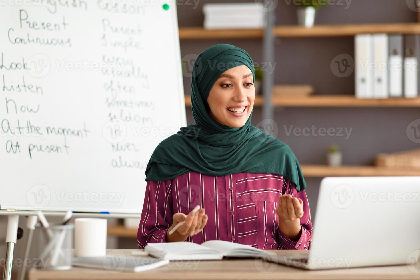 A young Middle Eastern teacher in hijab engages with students in an online English lesson. She appears enthusiastic and knowledgeable, with educational materials visible in the background. photo