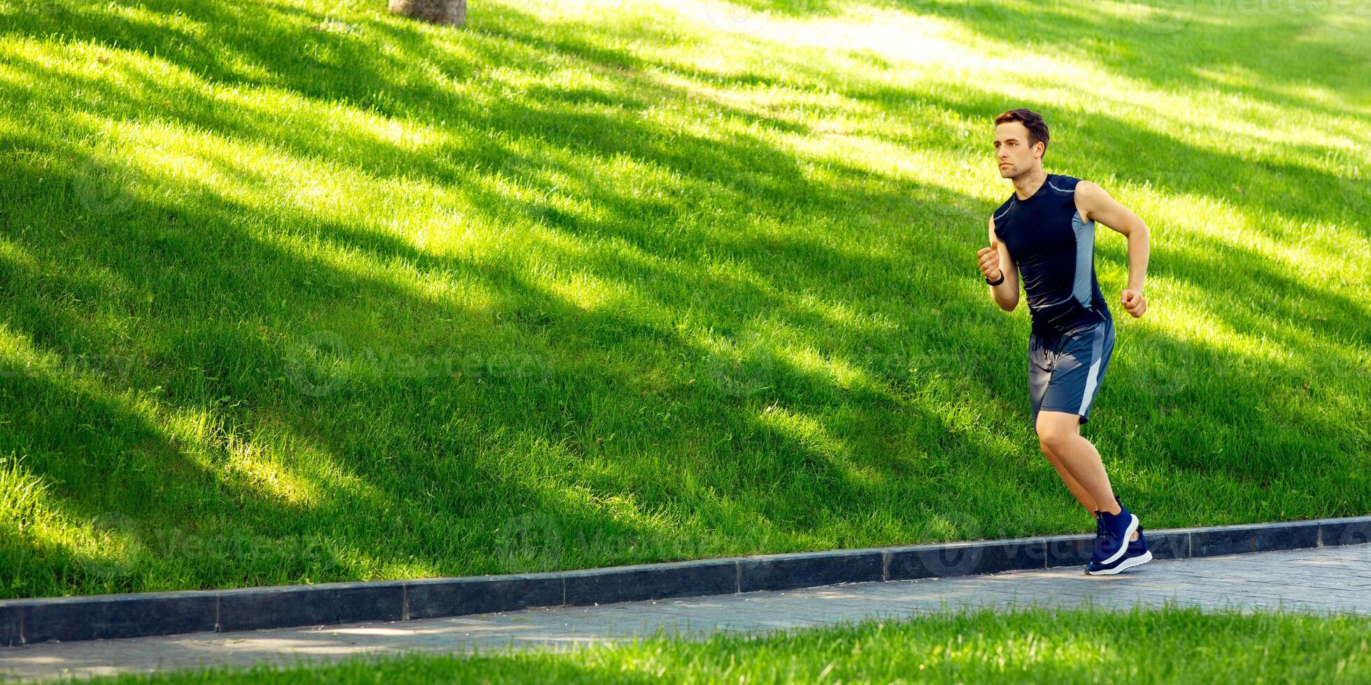 Young man runs at sunny day. Athlete in sportswear jogging in park with green grass, free space photo
