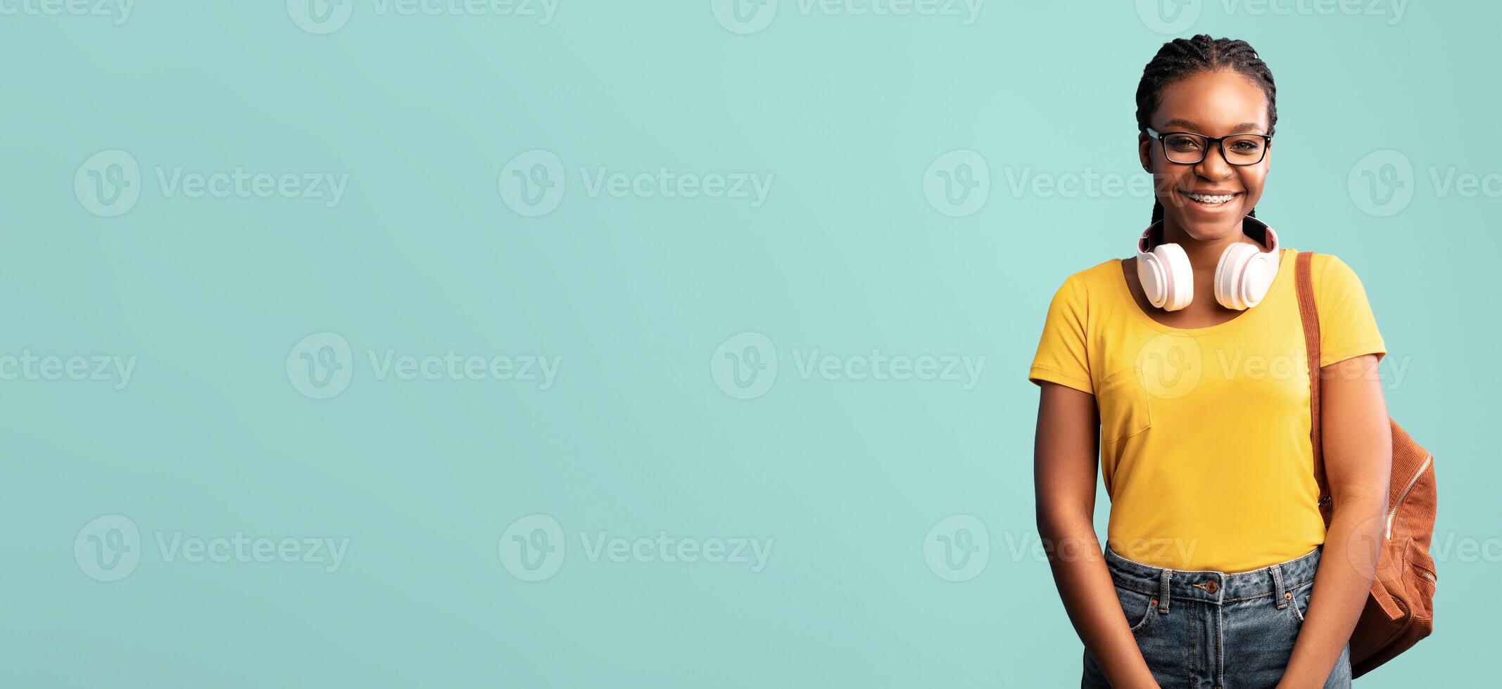 A young lady wearing a yellow shirt and glasses stands smiling with headphones around her neck and a backpack slung over one shoulder, set against a soft blue backdrop. photo