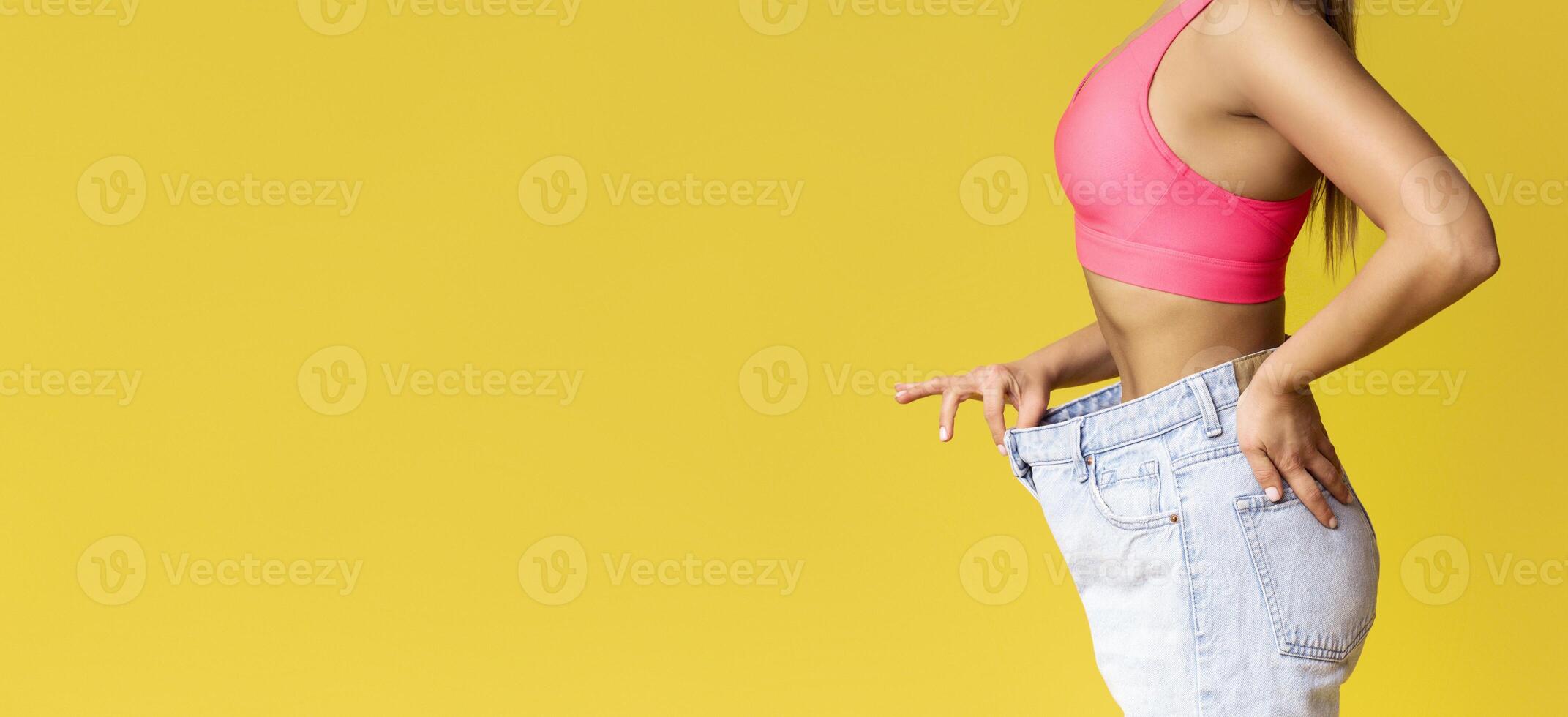 A woman stands with a joyful expression, showcasing a pair of large jeans while wearing a fitted pink top in front of a vibrant yellow background, highlighting her weight loss achievement, copy space photo