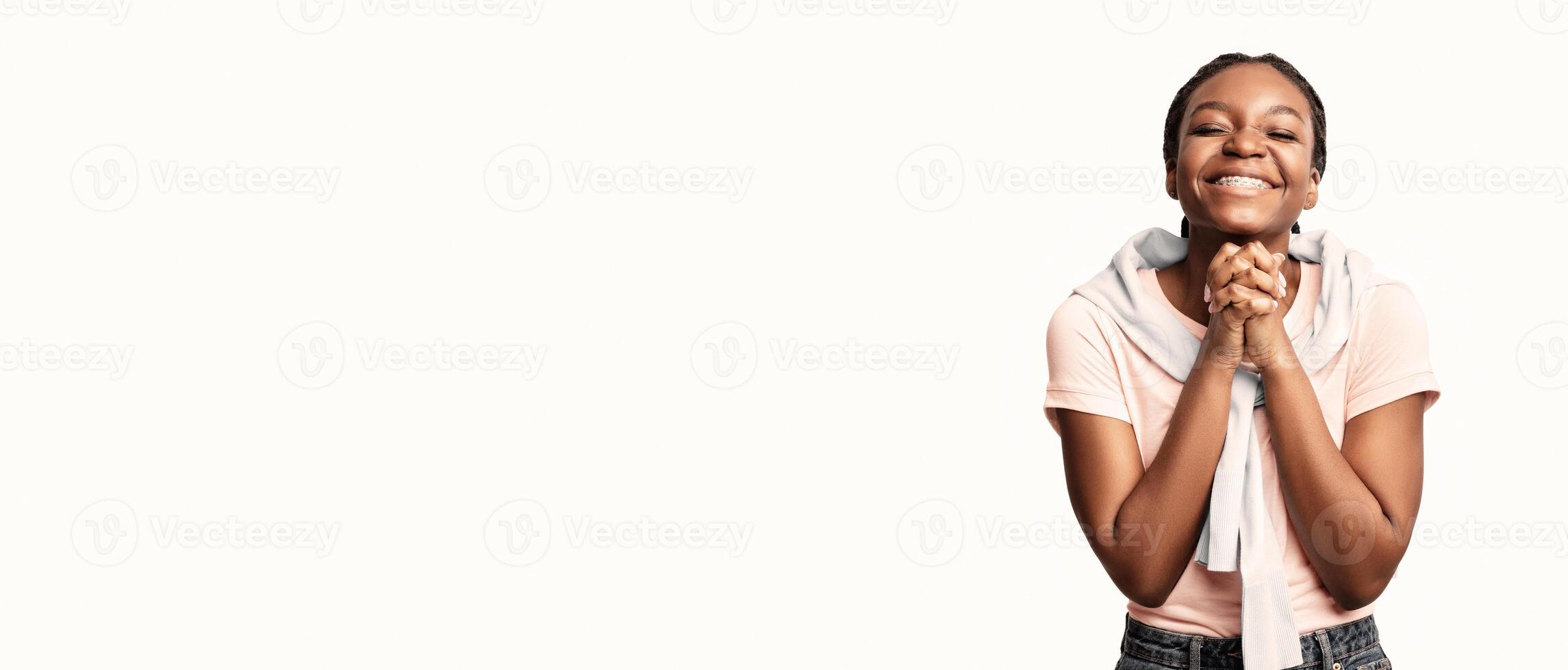 A cheerful young woman stands with her hands clasped together, smiling broadly in a bright, minimalistic environment. Her joyful expression conveys feelings of anticipation and gratitude. photo