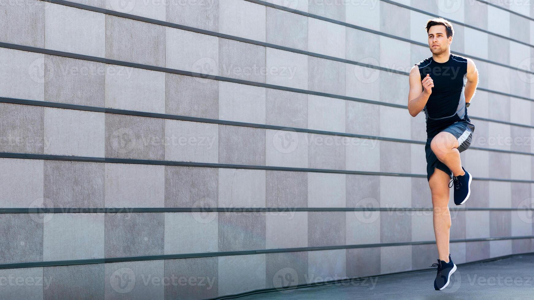 Jogging in street. Young man in sportswear and sneakers jumps and runs on gray wall background, free space photo