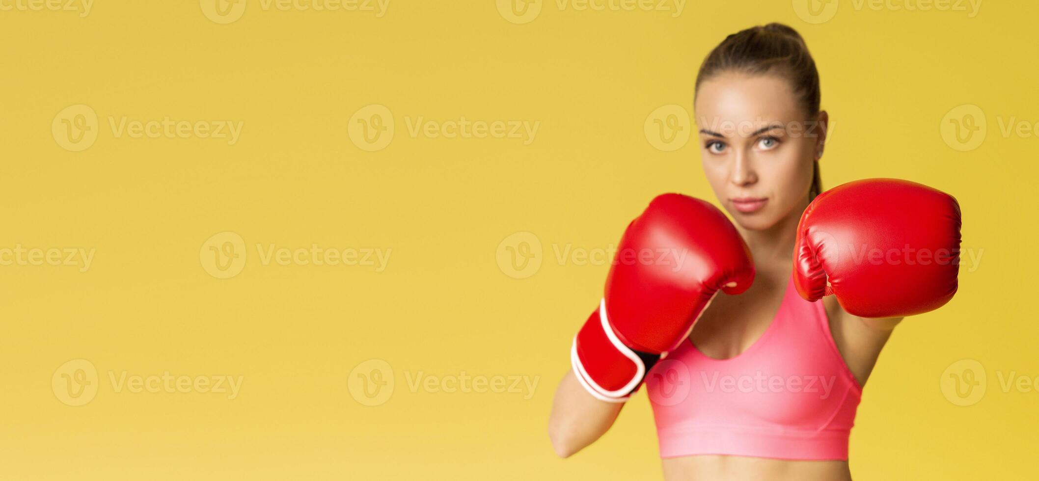 A determined woman in a pink athletic outfit wears bright red boxing gloves, showcasing her readiness for a match. The cheerful yellow background enhances her focused expression, copy space photo