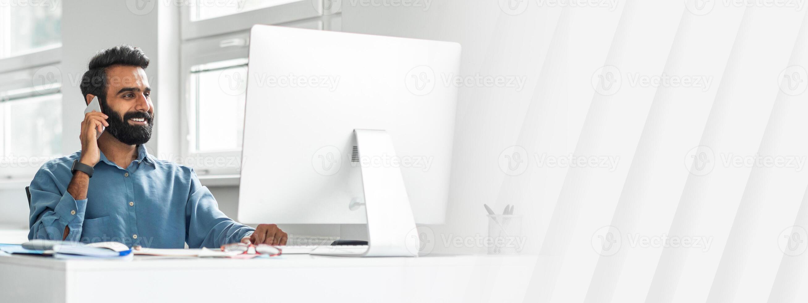 Handsome indian man office manager talking on cellphone at his workplace in office, sitting in front of computer, enjoying pleasant phone conversation, full length shot photo