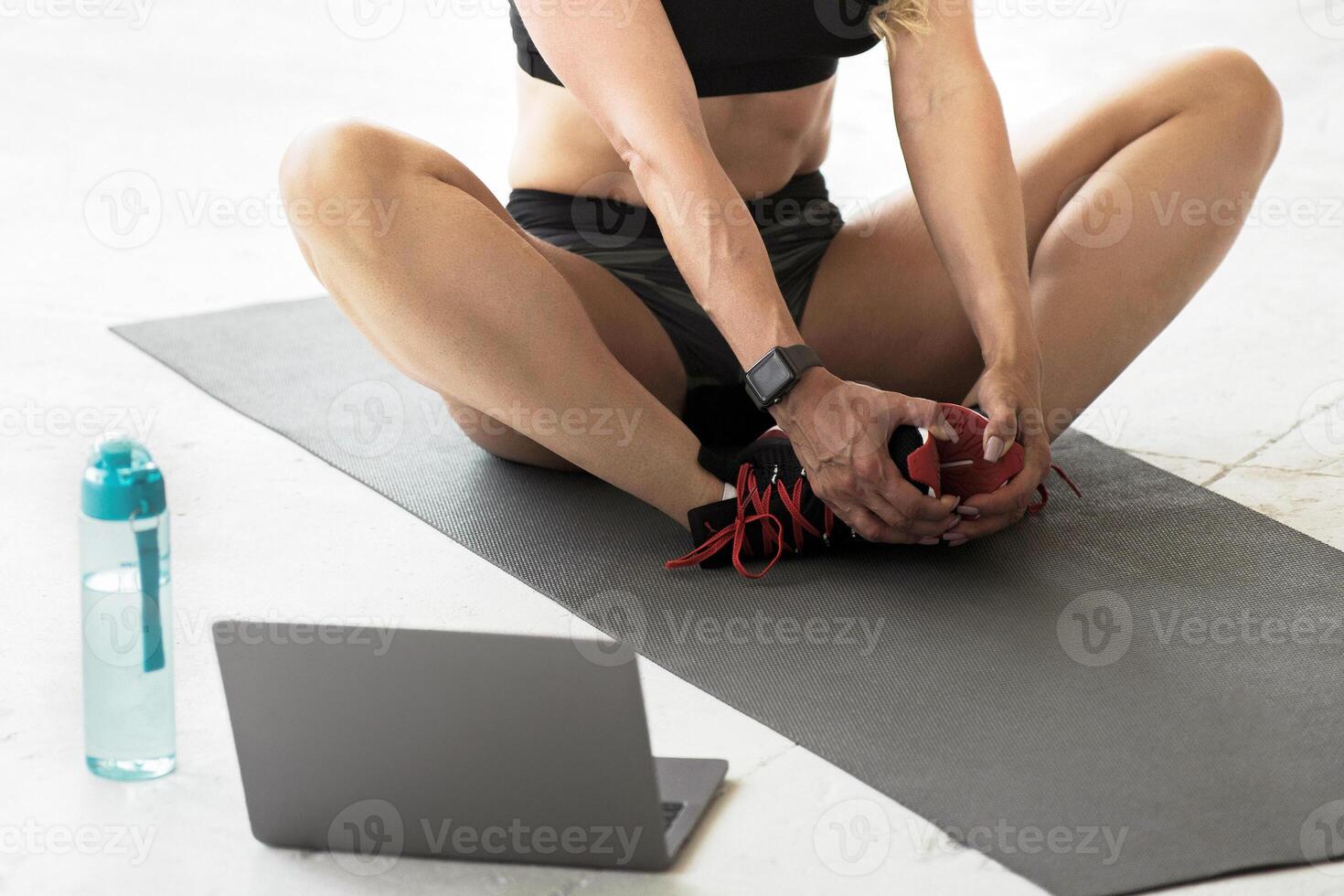 Online workout at home, streaming and lessons with coach remotely during lockdown. Muscular mature lady in sportswear sits on floor on mat with bottle of water and looks at laptop in loft photo