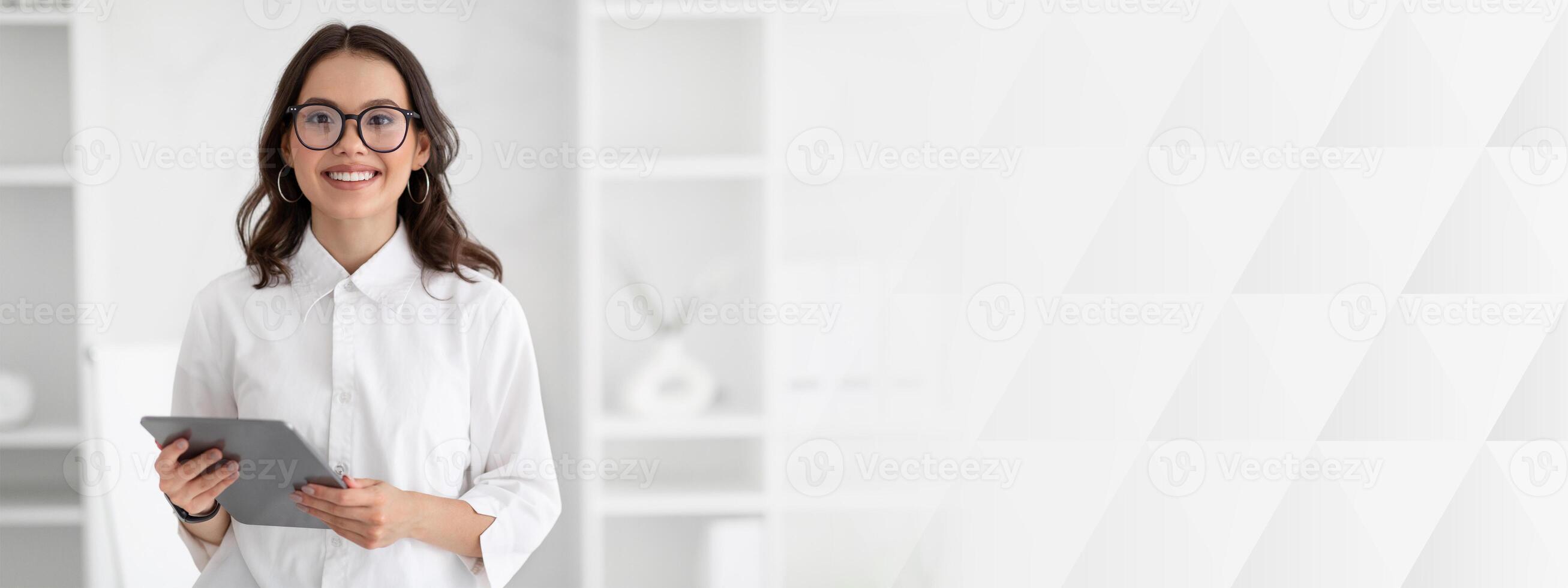 Happy millennial caucasian lady manager or teacher in white shirt and glasses uses tablet in light office interior. Modern app for business with device, work presentation, blog and study photo