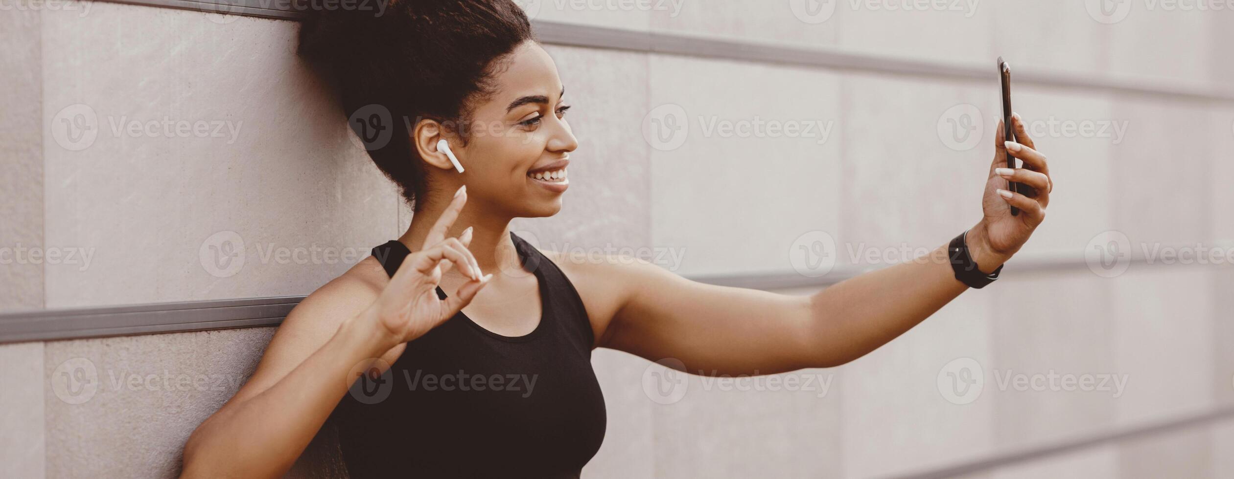 Selfie for sports blog. Muscular smiling african american girl in sport bra makes sign the peace and make photo on smartphone, panorama, free space