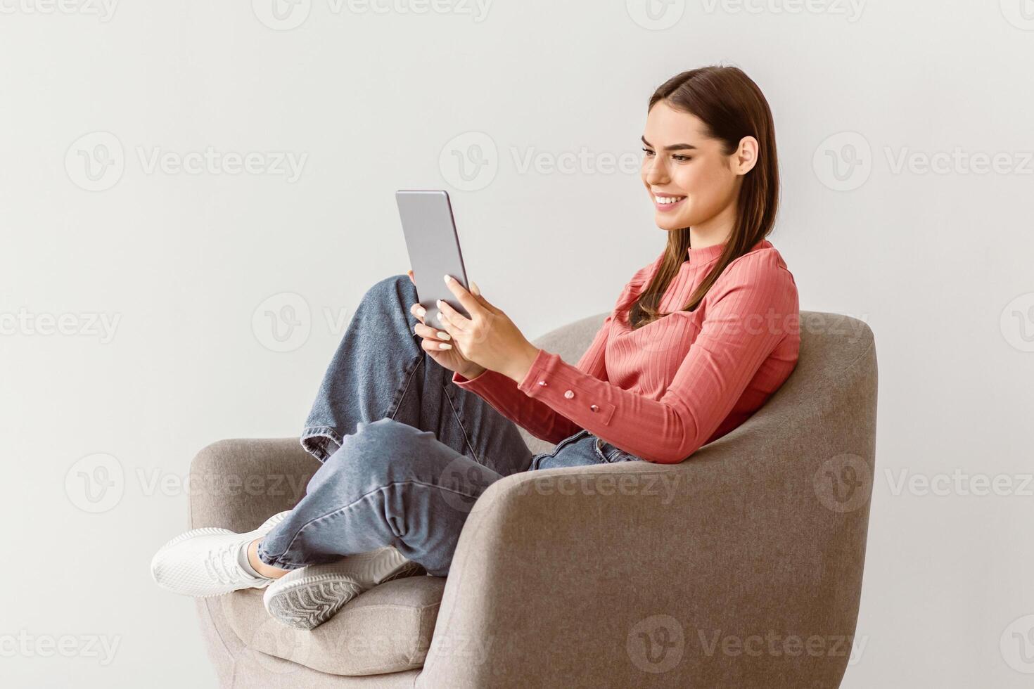 A young woman with brunette hair sits in a cozy armchair, engaged with her tablet. She wears casual attire and enjoys a peaceful moment, basking in the simplicity of her surroundings. photo