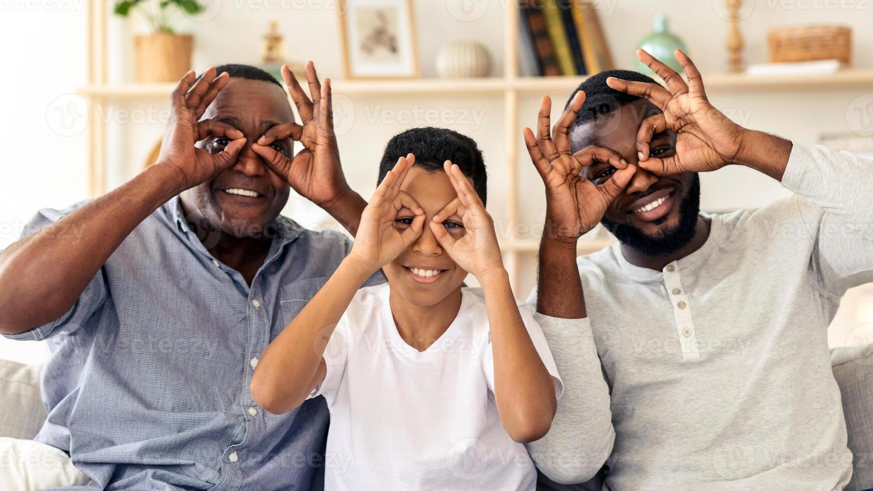 A grandfather, father, and son share a light-hearted moment indoors, each making funny hand gestures around their eyes, showcasing a bond filled with laughter and togetherness at home. photo