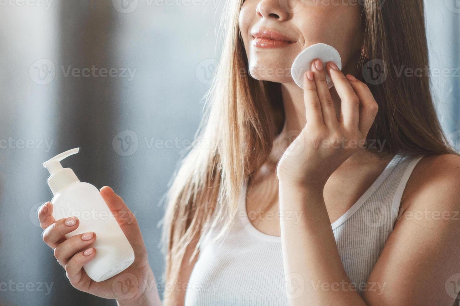 Skin care concept. Beautiful millennial woman removing makeup from her face near mirror at home photo
