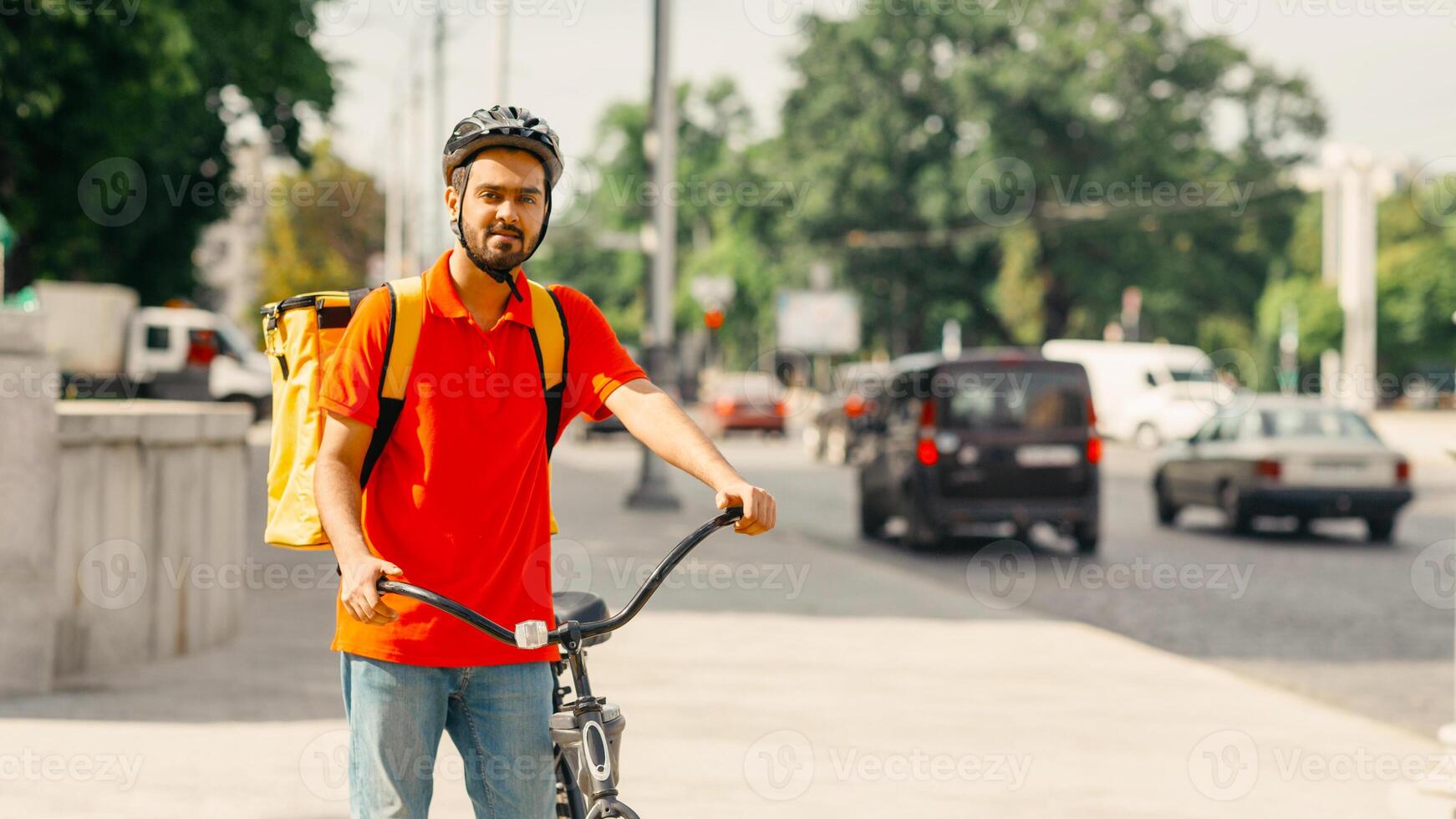Goods delivery service. Friendly courier in safety helmet with big bag, stands with bike at crossroads in city, copy space photo