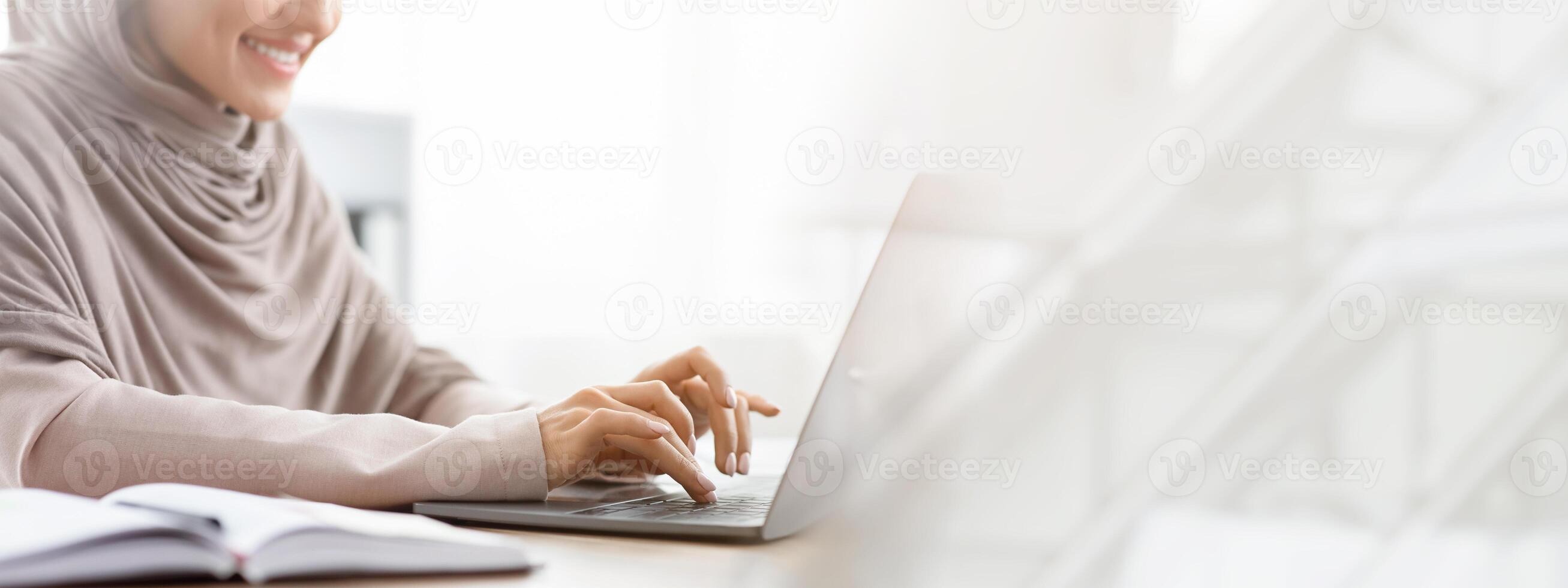 Unrecognizable muslim woman in hijab working on laptop computer in office, typing on keyboard and smiling, cropped image with empty space photo