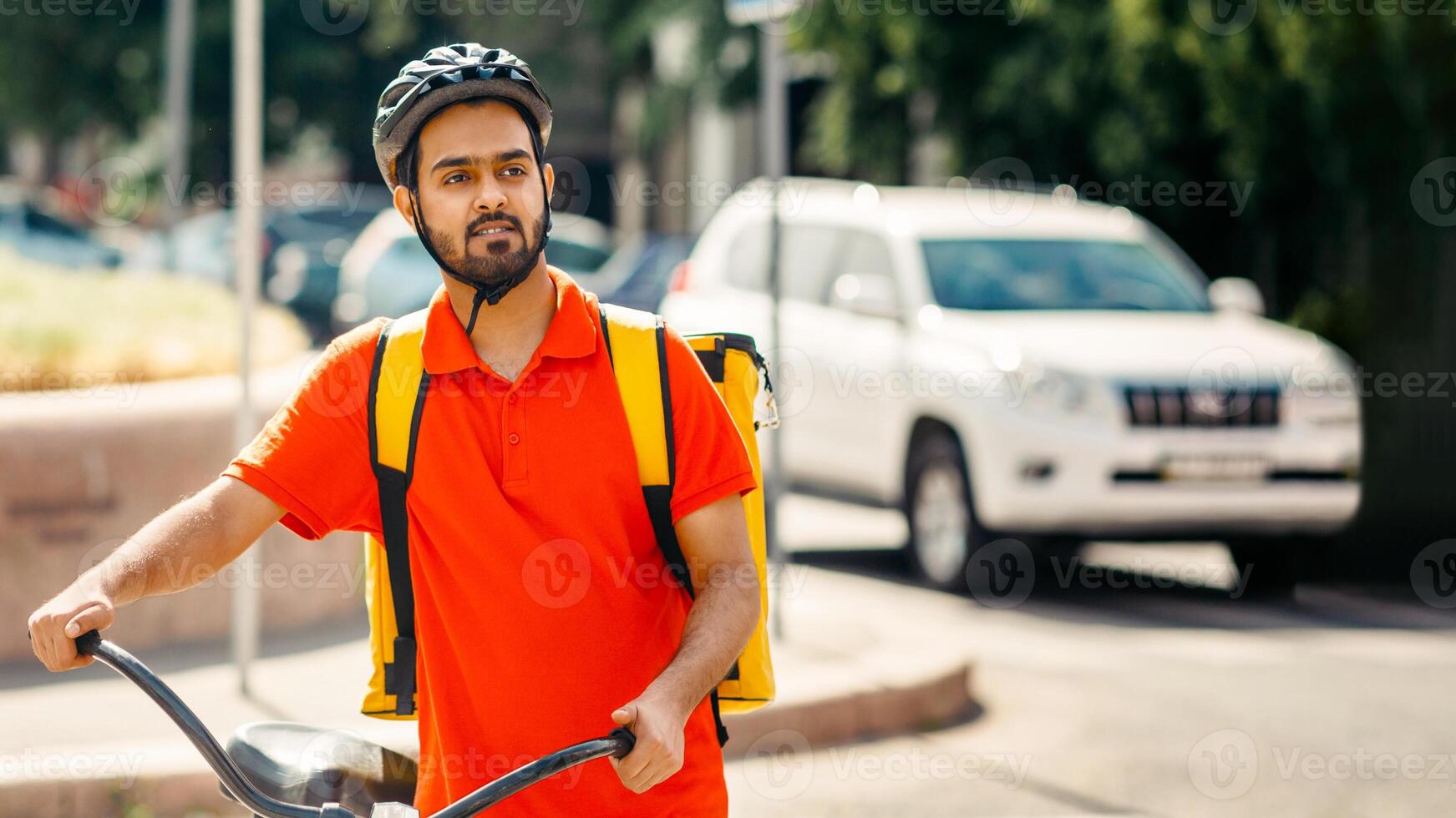 Mobile work in city, fast delivery and sporty lifestyle. Attractive millennial arabian guy courier in orange t-shirt and protective helmet, with big backpack and bicycle looks for address of client photo