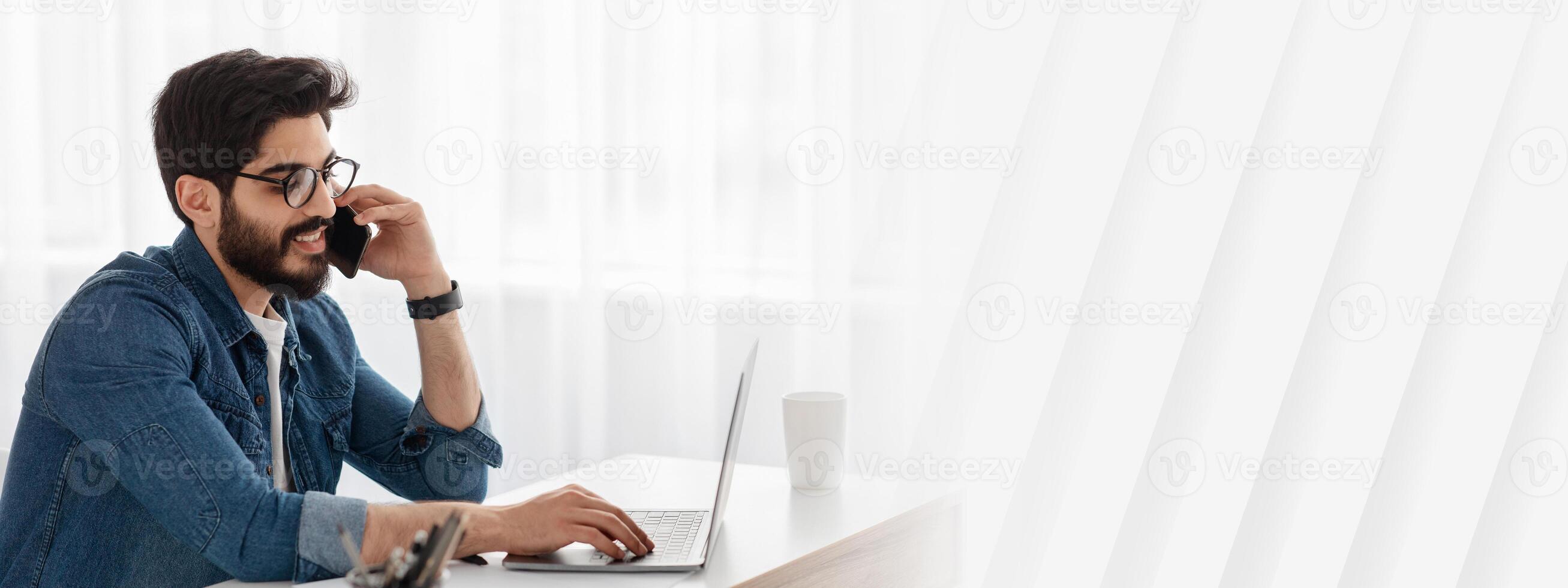 Mobile business communication and entrepreneurship. Arab businessman talking on cellphone and using laptop computer, sitting at workplace at home office, empty space photo