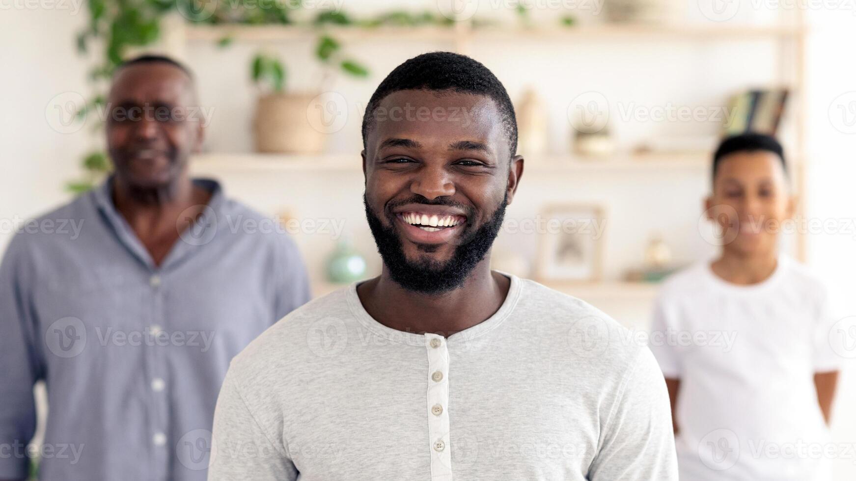 A cheerful man in a light grey shirt smiles widely in a well-lit room. Family members stand behind him, creating a warm and inviting atmosphere filled with plants and decor. photo