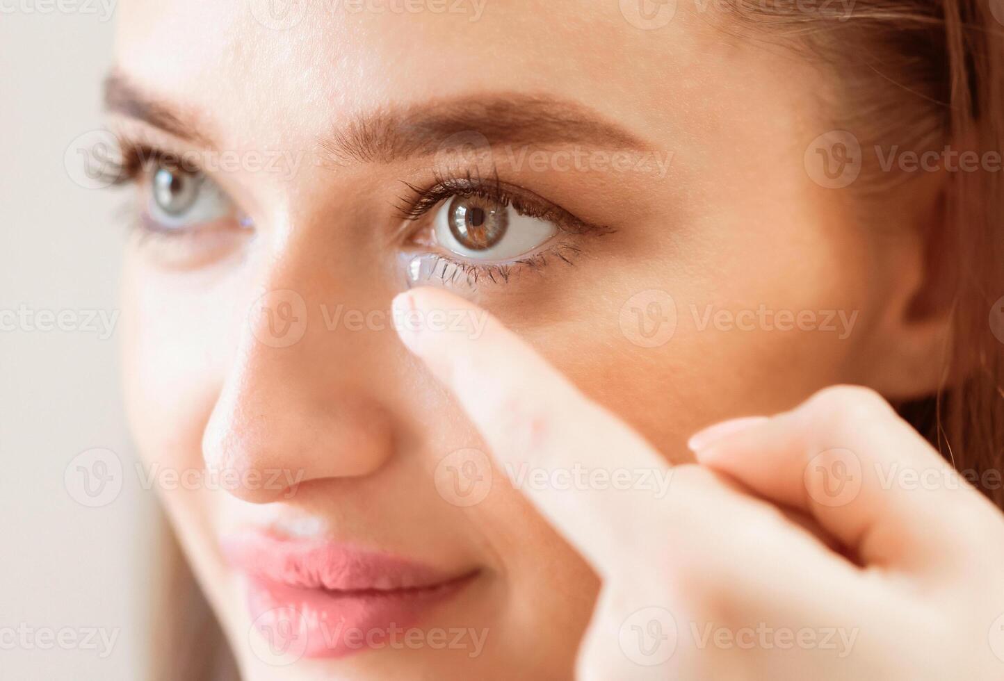 A young woman carefully places a contact lens in her right eye, focusing intently. The soft lighting highlights her features as she maintains a steady hand and calm demeanor. photo