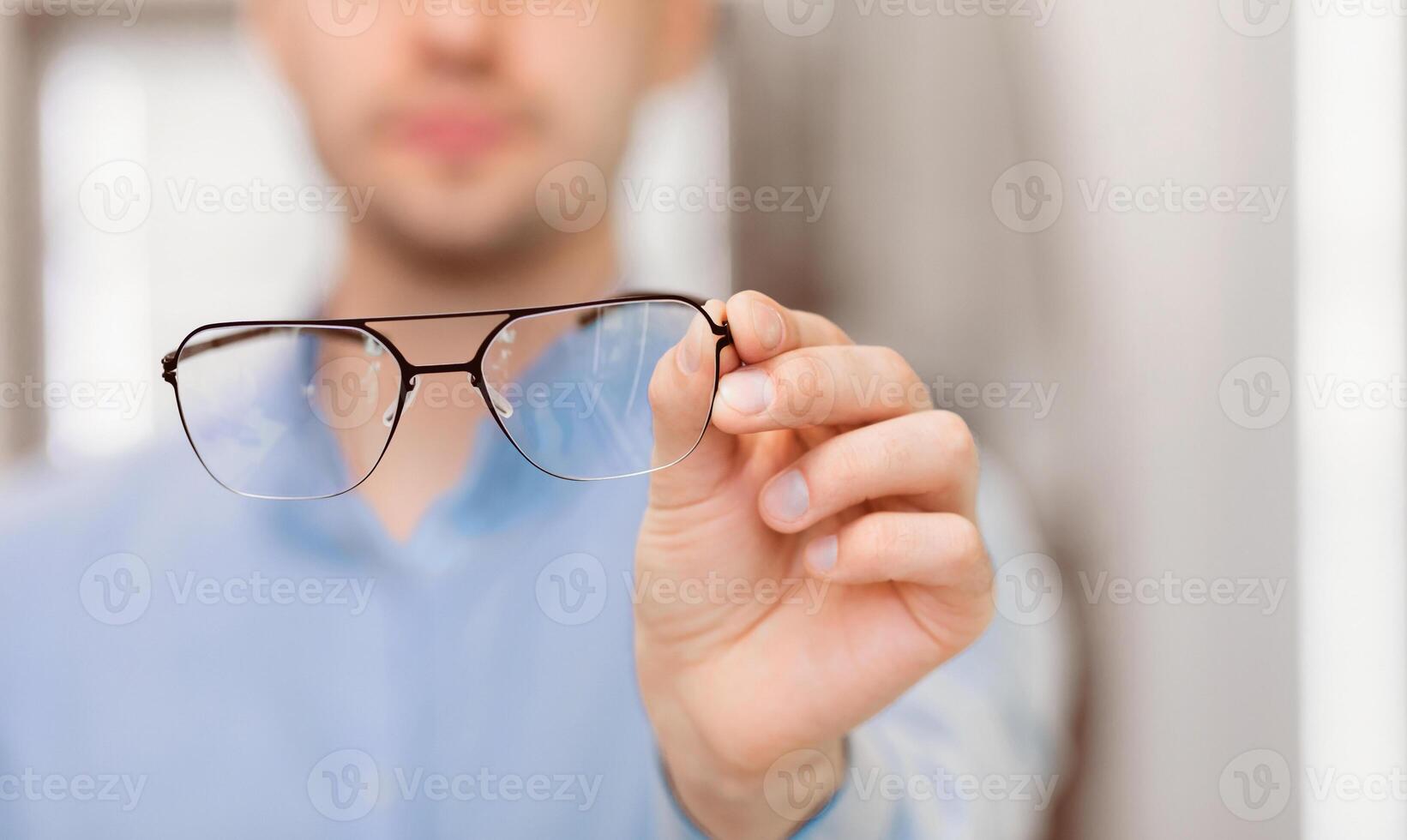 A person is holding a pair of eyeglasses in front of them in a bright indoor space. The focus is on the glasses, showcasing their design, while the person remains slightly blurred in the background. photo