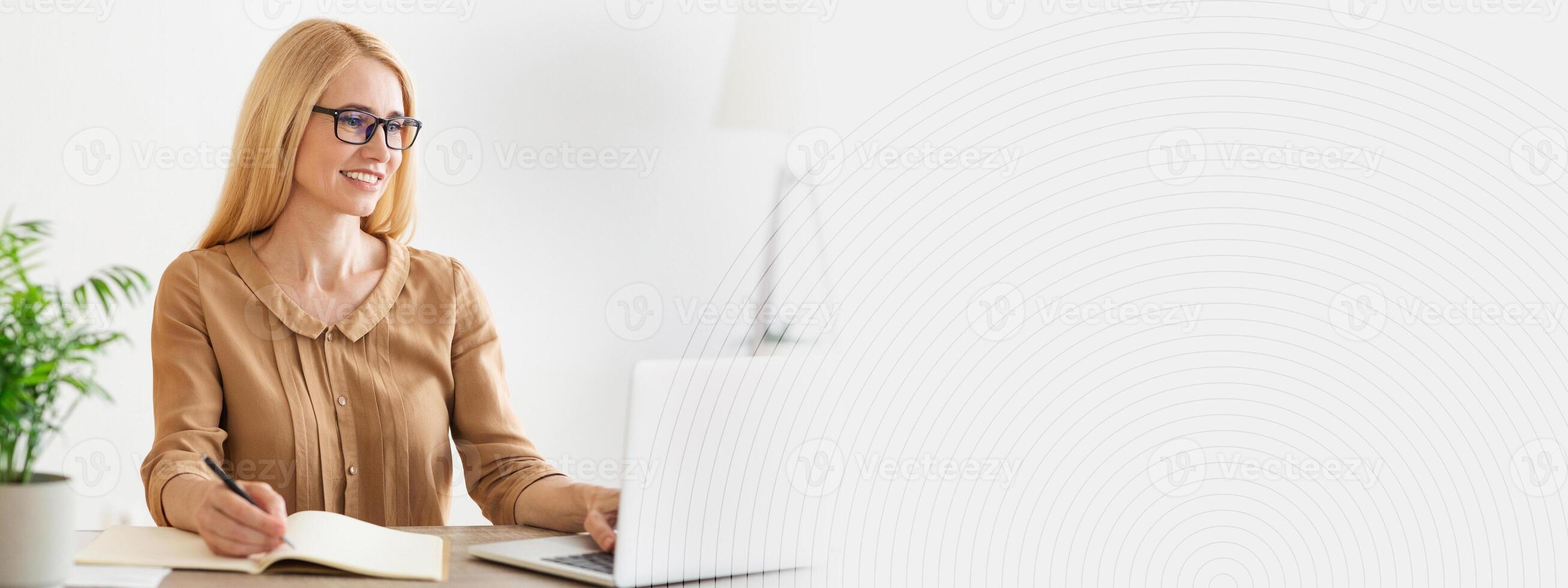 A woman sits at a light, airy workspace, focused on her laptop while taking notes in a notebook. A potted plant adds a touch of greenery, enhancing the cozy atmosphere, copy space photo
