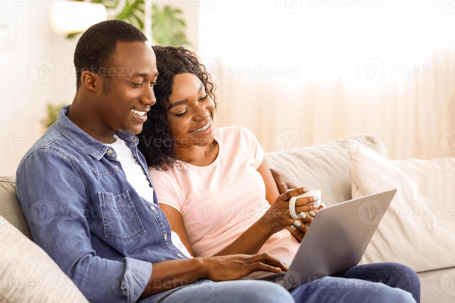 Happy black couple using laptop at home, looking for new destination to travel, copy space. African american man and woman in love sitting on couch at living room, drinking tea and websurfing photo