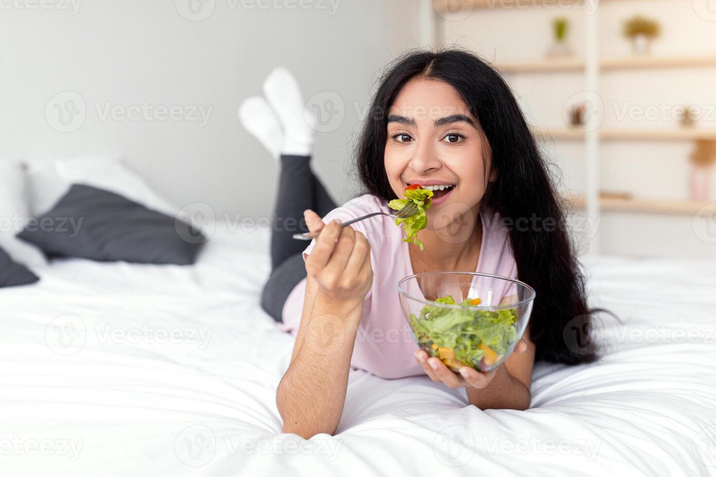 A millennial Indian woman lies on her bed, cheerfully eating a fresh salad from a bowl. She embraces a healthy lifestyle by incorporating nutritious meals into her daily routine. photo