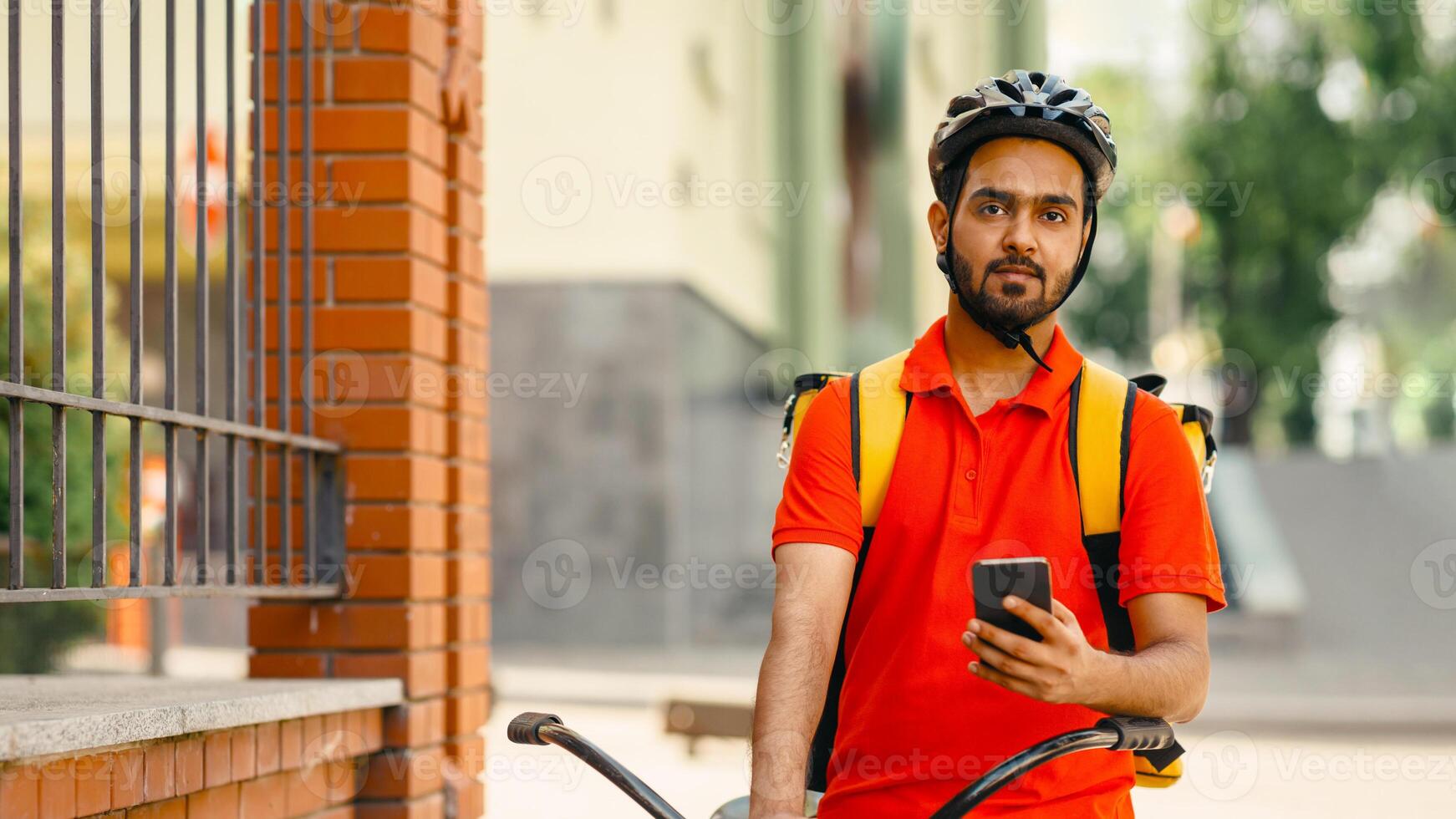 Home delivery service. Friendly courier in protective helmet, with yellow backpack, sits on bicycle and holds smartphone in hand on street, copy space photo