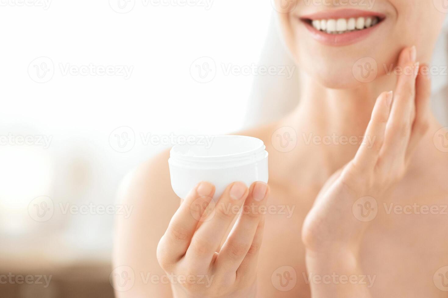 Face care concept. Cropped of unrecognizable smiling woman wrapped in towel using cream. Jar with face cream in young lady hands, applying moisturizer on face after shower in morning at home photo
