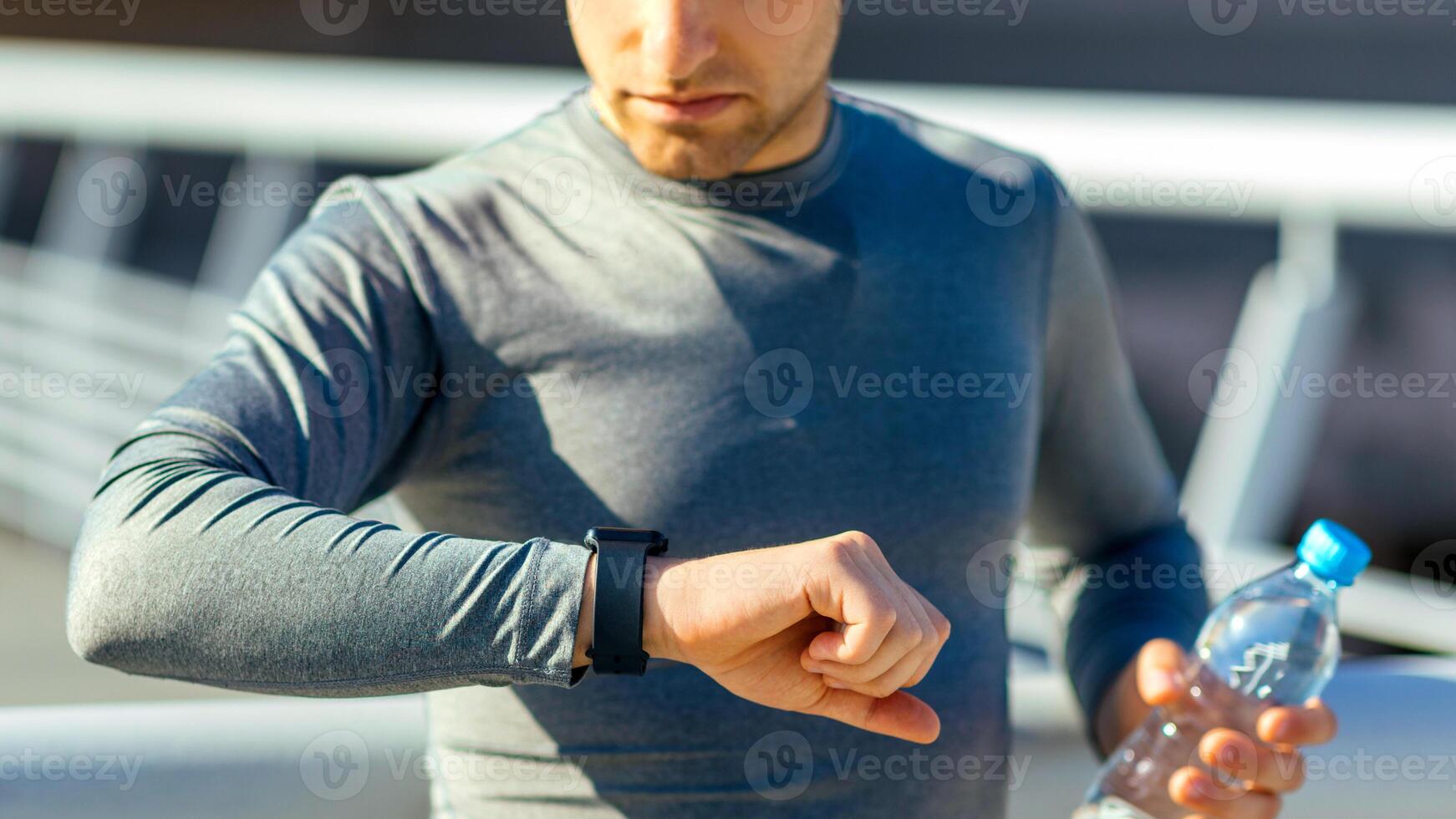 Time for training. Concentrated young man in wireless headphone with bottle of water looks at fitness tracker at stadium, panorama, free space photo