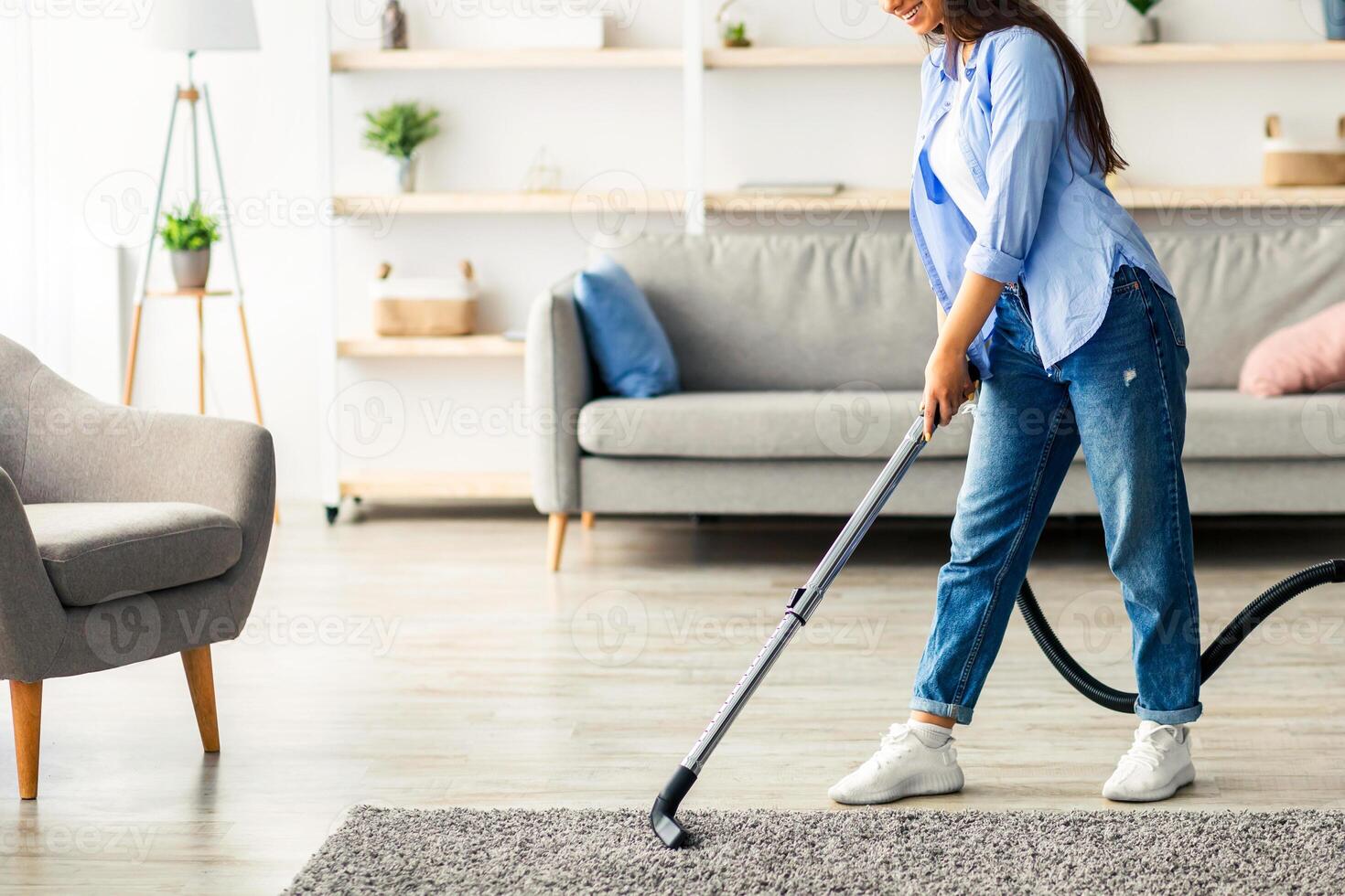 A young woman enjoys her time cleaning the carpet in her modern living room. She uses a vacuum cleaner while wearing casual clothes, with sunlight streaming through the window, copy space photo