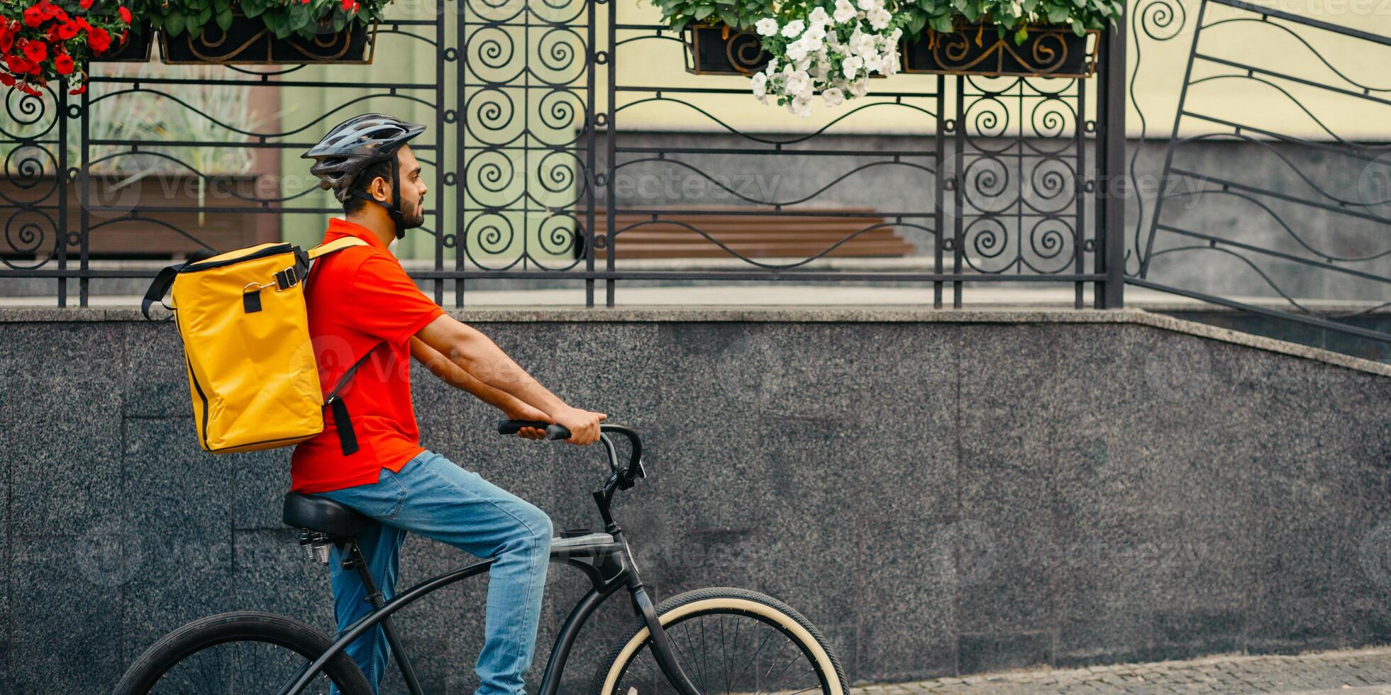 Courier carry out orders for delivery of goods. Man in helmet and big yellow backpack rides bicycle at city at summer, side view, free space photo