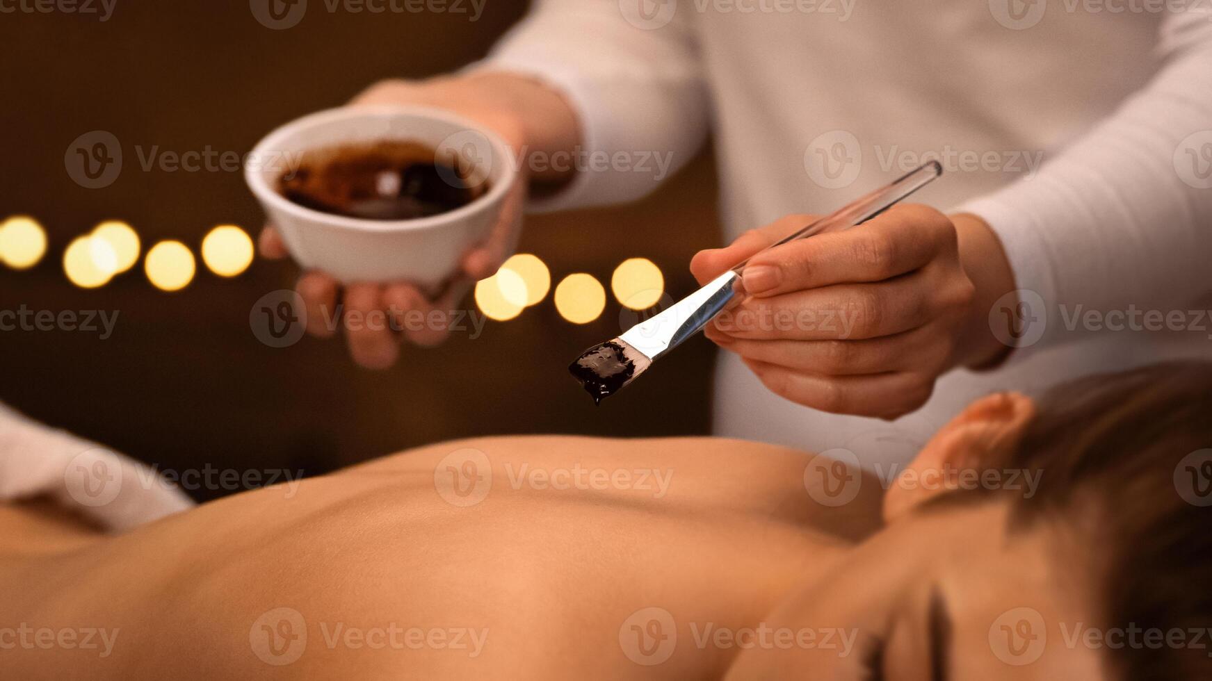 Unrecognizable massage therapist applying exfoliating body mask with brush on relaxed woman back at spa, body care concept. Smiling young lady having body treatment with aroma at modern spa, closeup photo