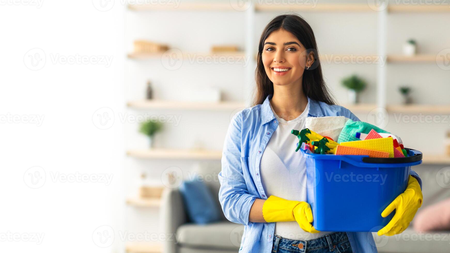 A young woman is happily holding a blue container filled with cleaning supplies while smiling. She is dressed casually and appears to be cleaning her home space during the day, copy space photo