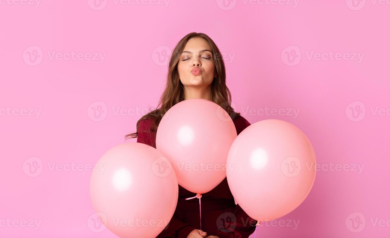 A young woman makes a playful kissy face while holding three soft pink balloons, showcasing a cheerful and lighthearted atmosphere in a bright pastel setting. photo