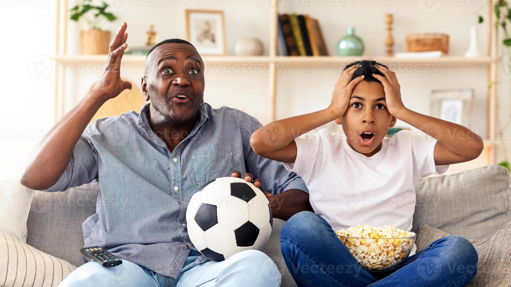 A man and a boy sit on a couch, transformed by the thrill of a soccer game. The boy looks shocked, while the man expresses enthusiasm, holding a soccer ball beside him. photo