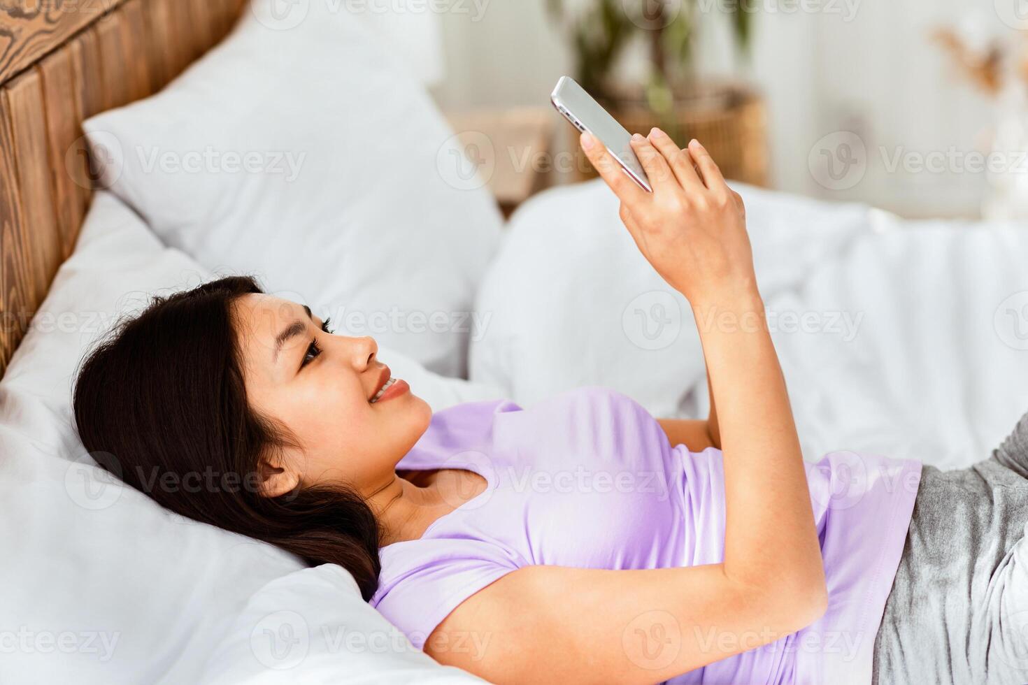 Happy Korean Girl Using Cellphone Texting In Online Chat App Lying In Bed At Home On Weekend In The Morning. photo