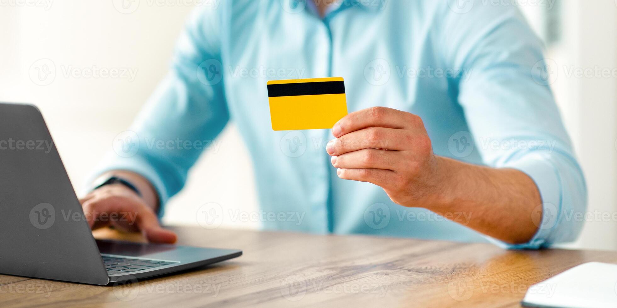A young Arab entrepreneur sits at a wooden desk, actively using a laptop and holding a credit card, focused on a business task in a modern office environment. photo
