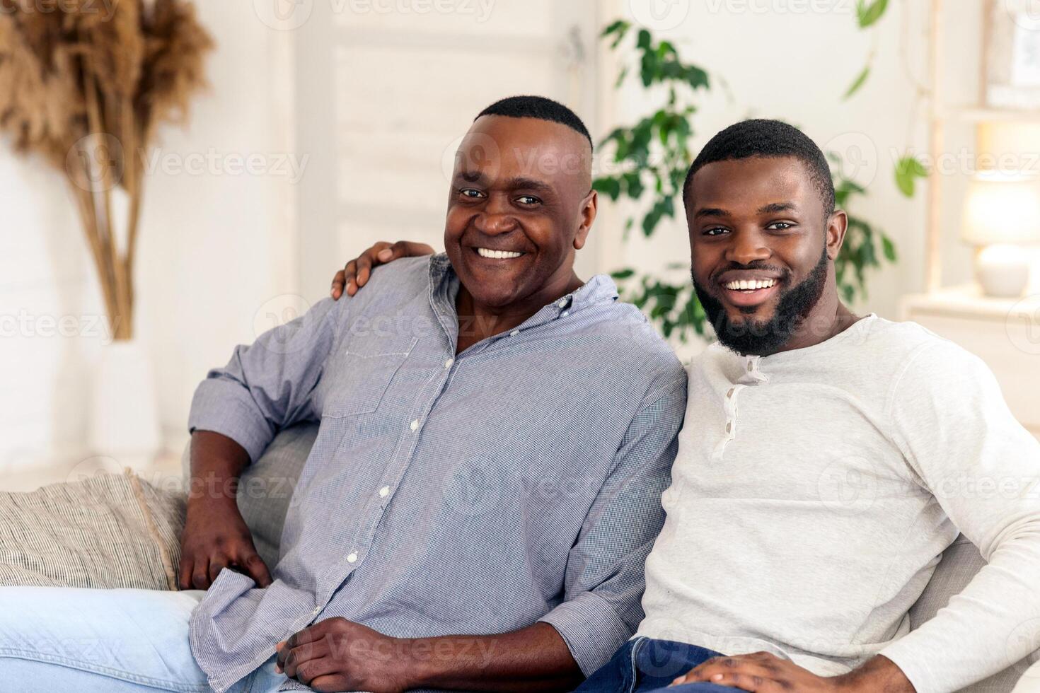 A father and son share a joyful moment together in their cozy living room, smiling warmly as they relax on the sofa surrounded by plants and comfortable decor, creating a heartwarming atmosphere. photo
