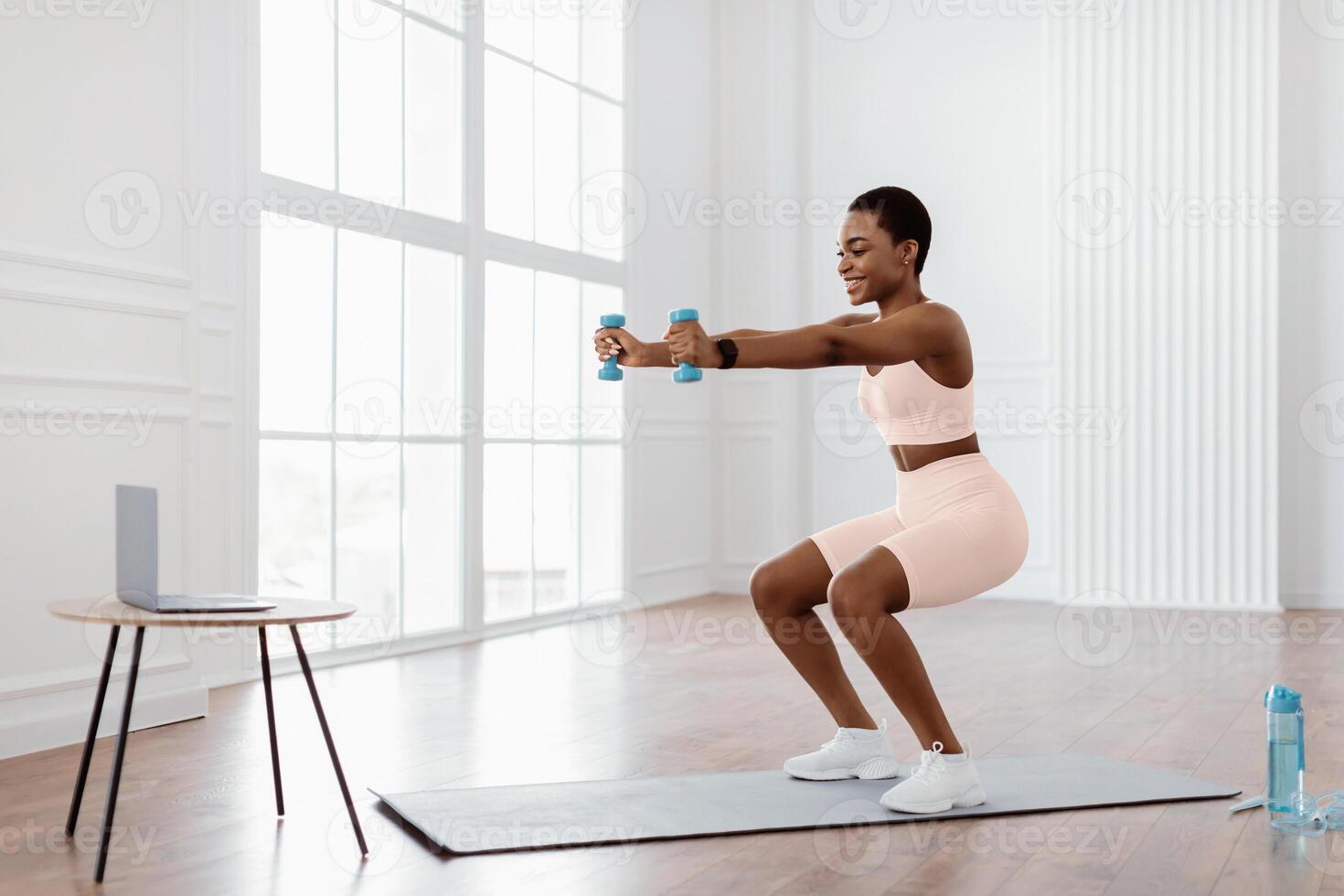 A young African American woman exercises at home, performing squats while holding dumbbells. She follows an online fitness routine, promoting a healthy and active lifestyle. photo