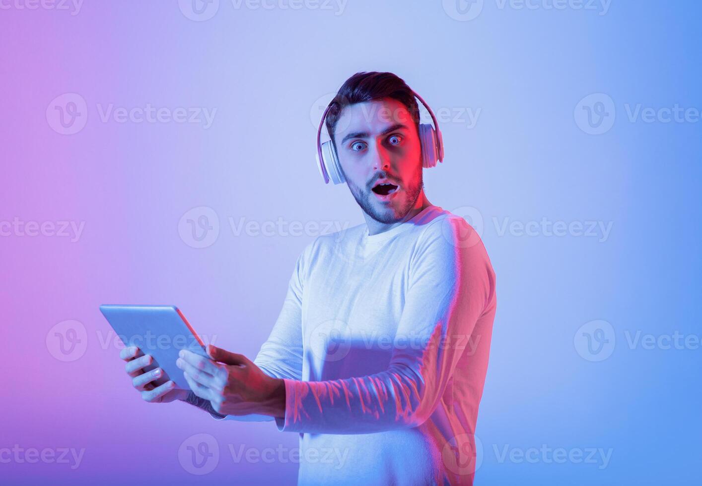 A young man wearing headphones expresses surprise while holding a tablet. The colorful background enhances the excitement of the moment, creating a lively atmosphere. photo