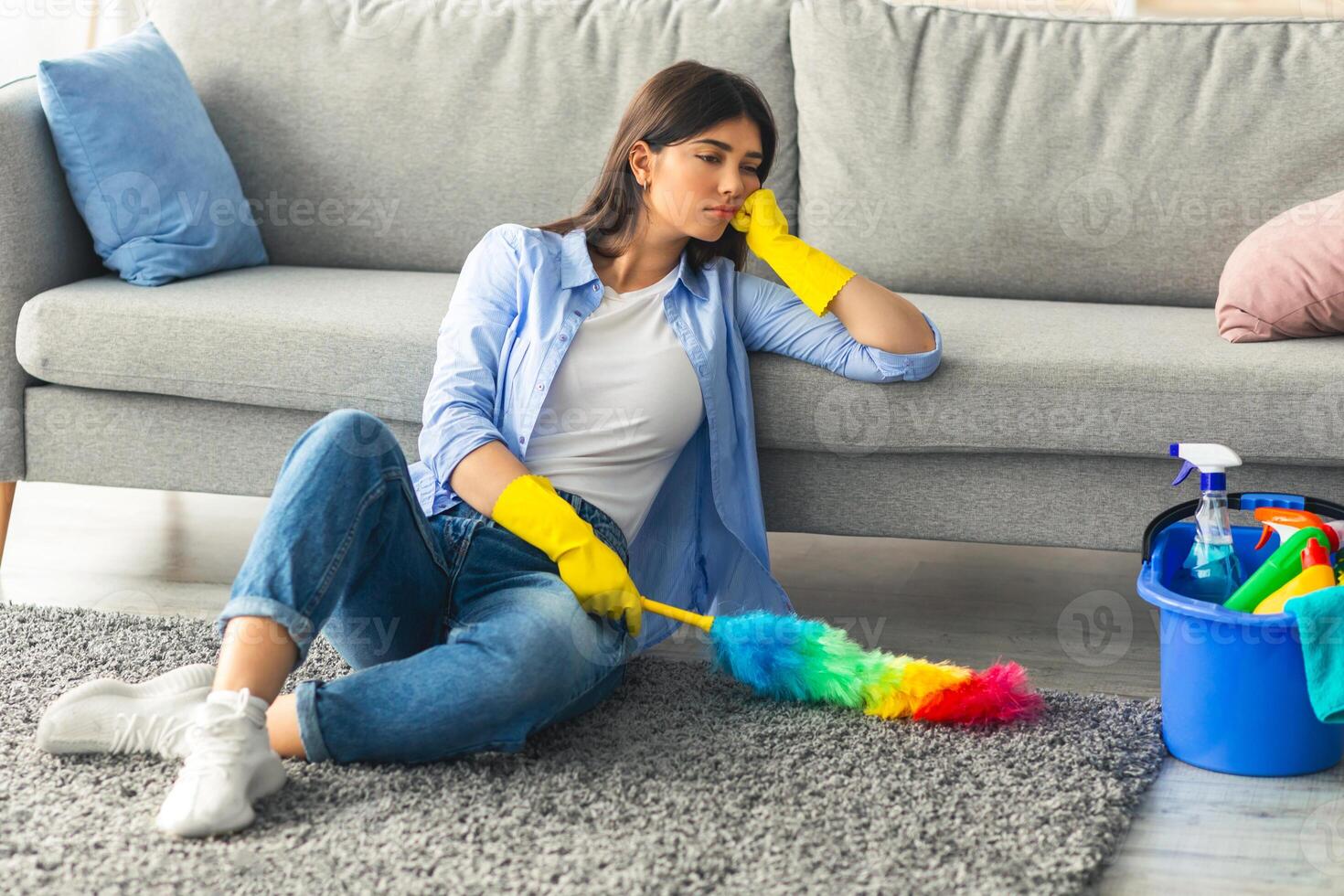 A young woman in casual attire sits on the floor of her living room, wearing yellow gloves. She looks bored while dusting the carpet with a colorful duster. Cleaning supplies are nearby. photo