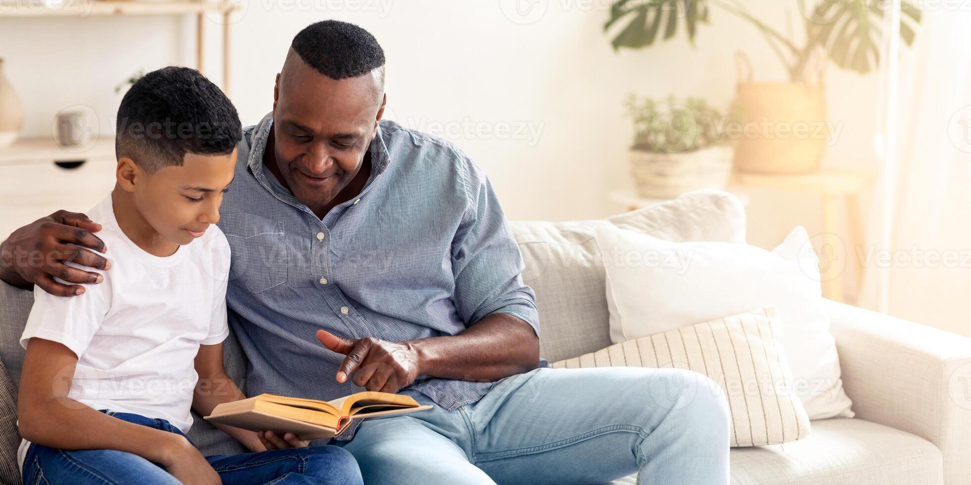 A grandfather and his young grandson sit together on a comfortable sofa, sharing a moment as they read a book. The warm sunlight filters through the window, creating a cozy atmosphere. photo